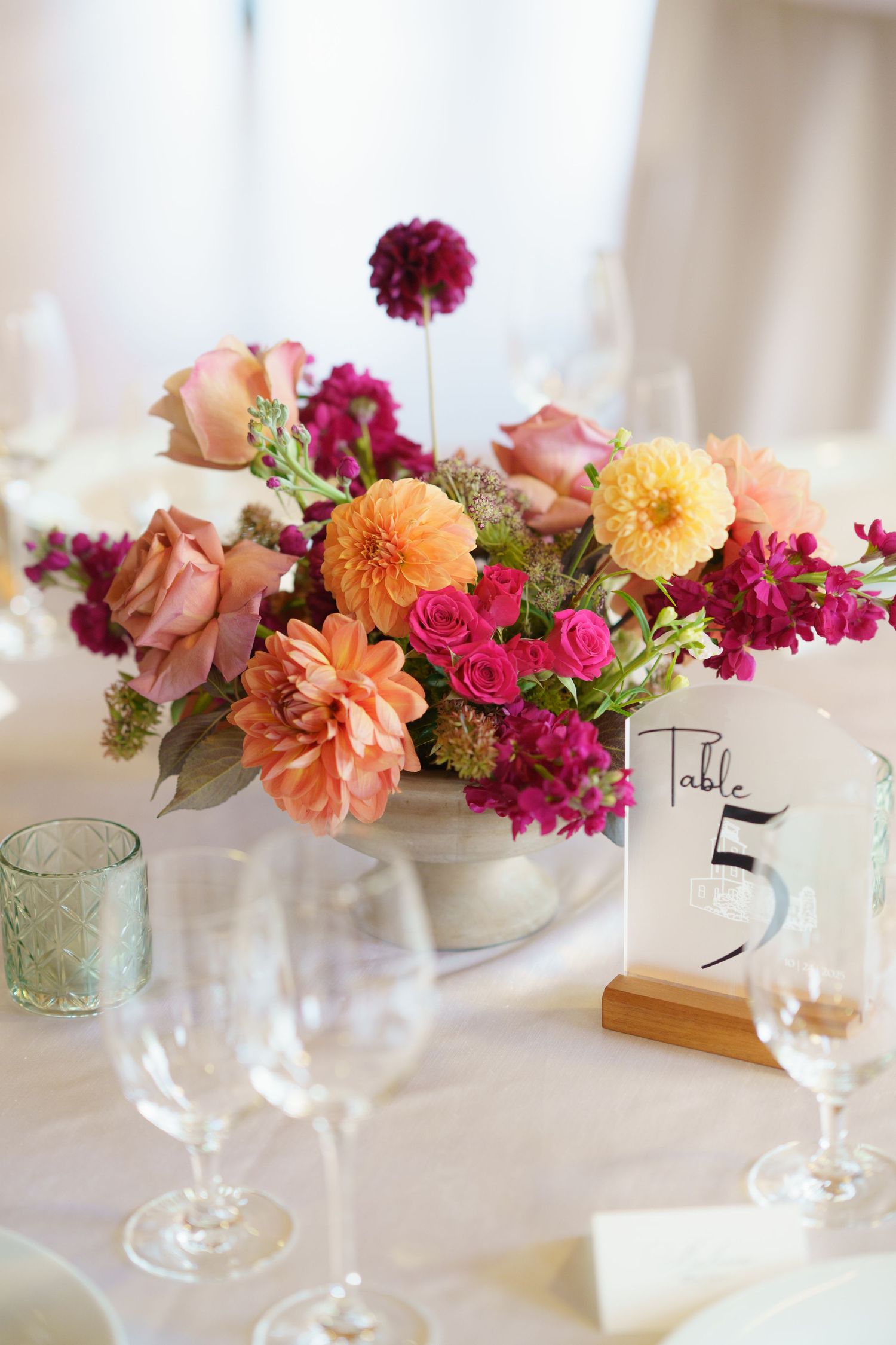 Vibrant wedding centerpiece with orange dahlias, pink roses, and magenta blooms in a white bowl on a reception table.