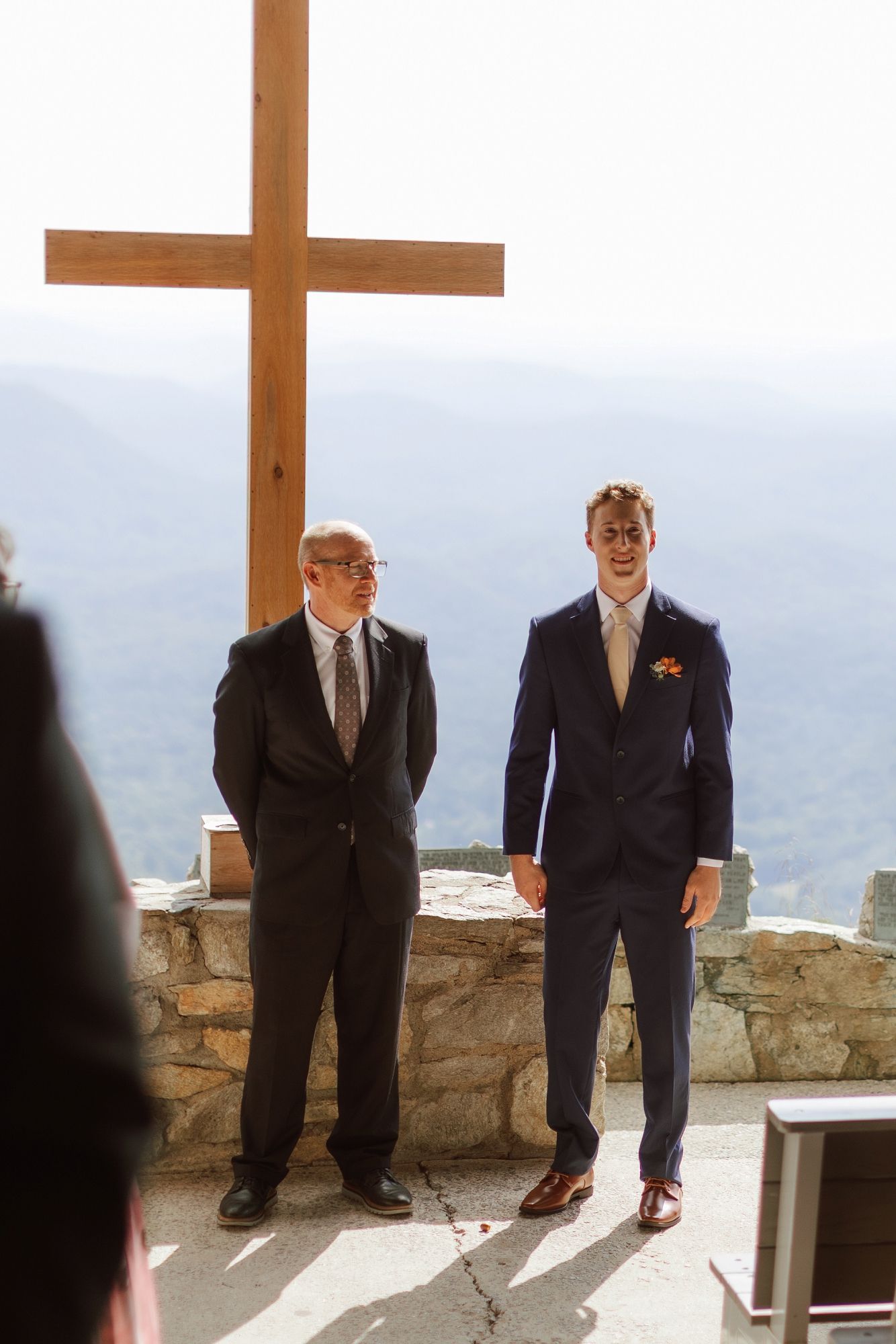 Groom in navy suit smiling at wedding altar with large wooden cross and mountain views backdrop.