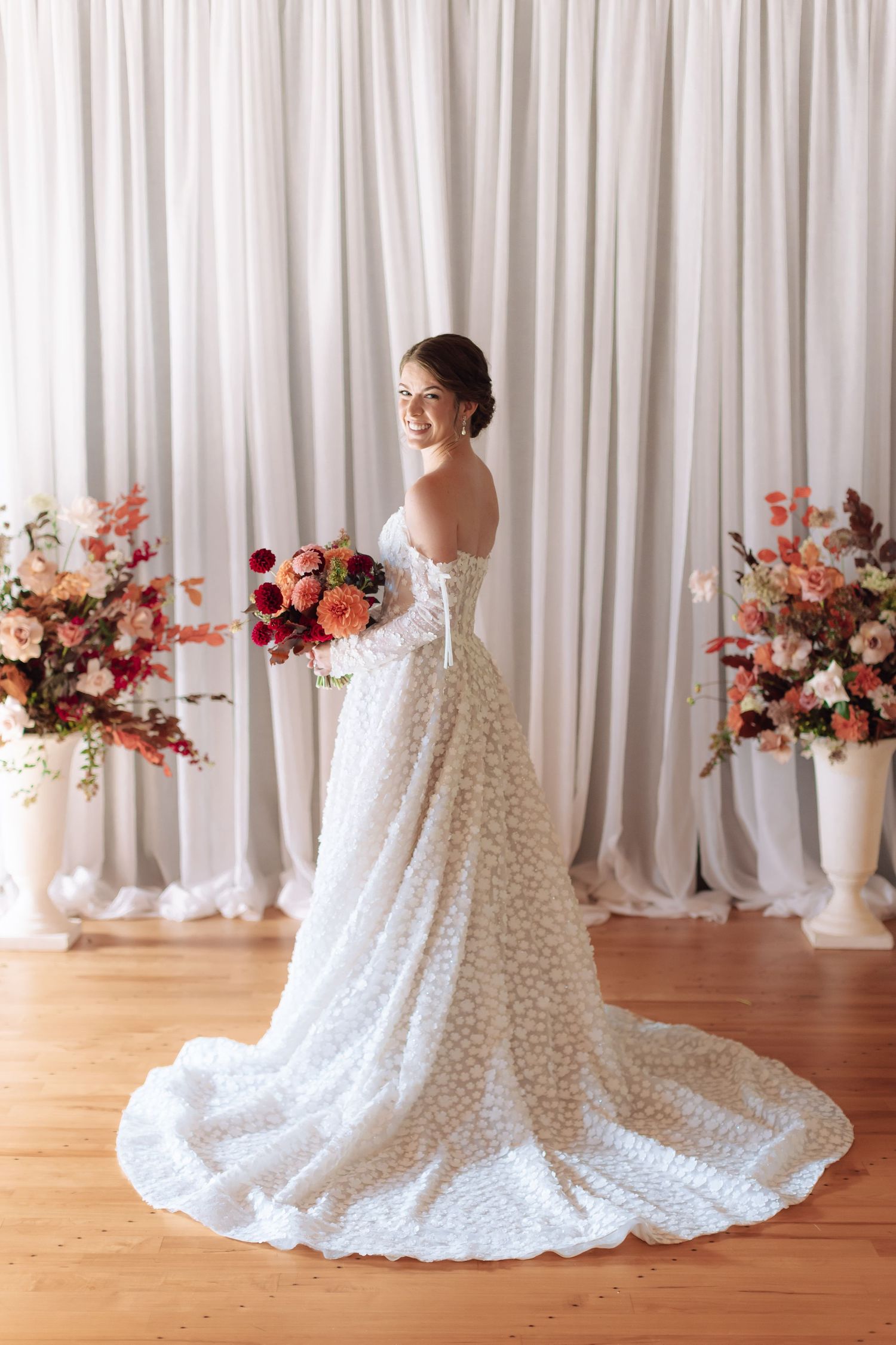 Smiling bride in a white lace gown with train holds a bouquet, posing before draped curtains and floral arrangements.