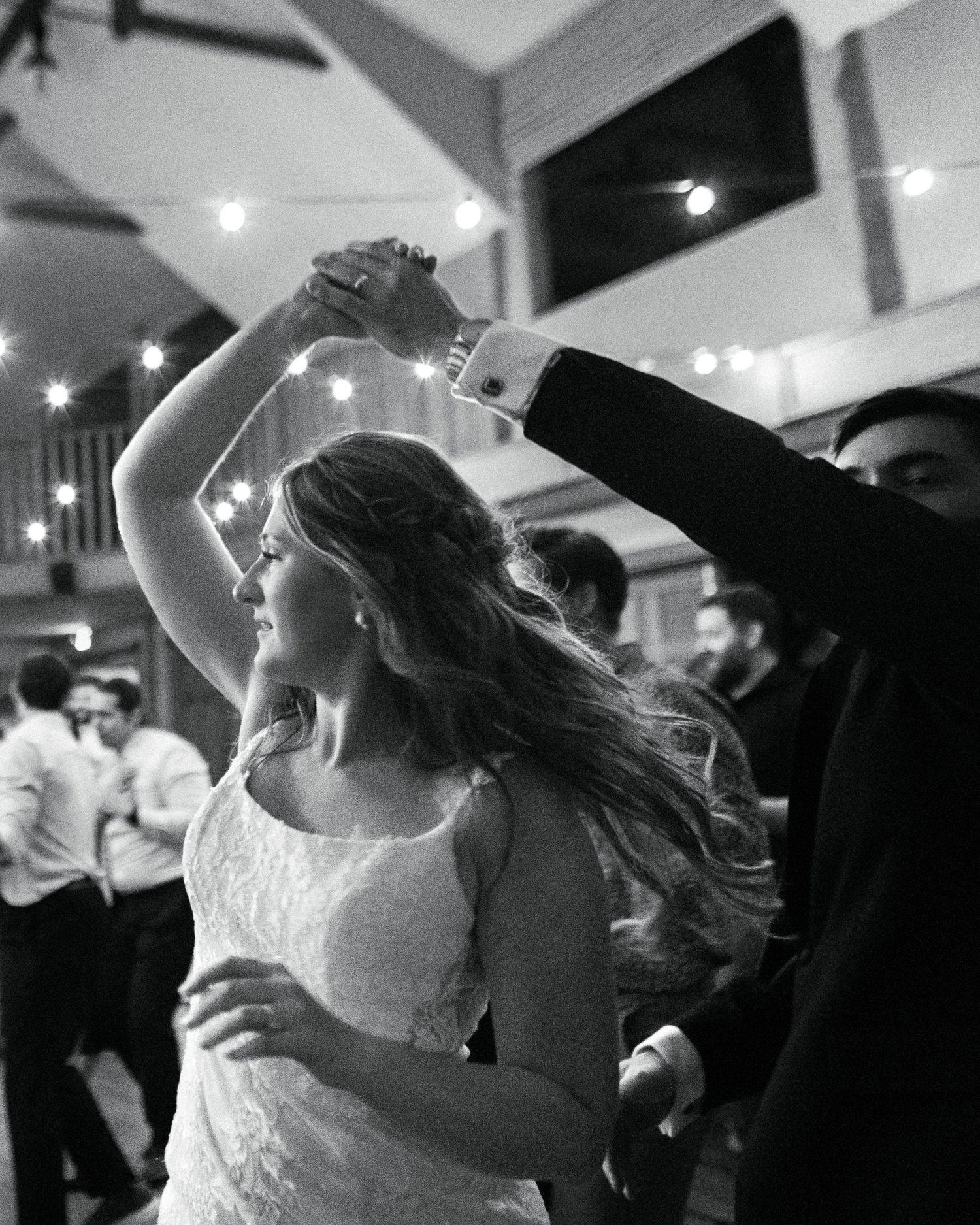 Black and white image of bride dancing at a wedding reception with twirling motion and string lights in the background.