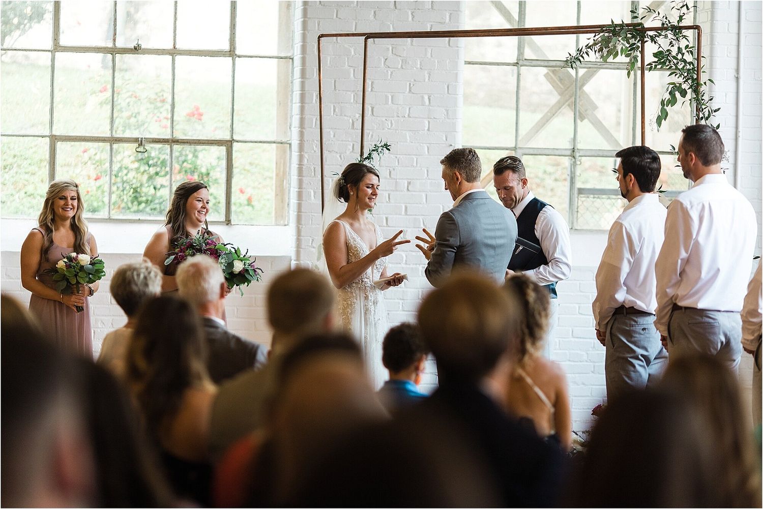 Wedding ceremony with copper arch backdrop in bright industrial space with large windows.