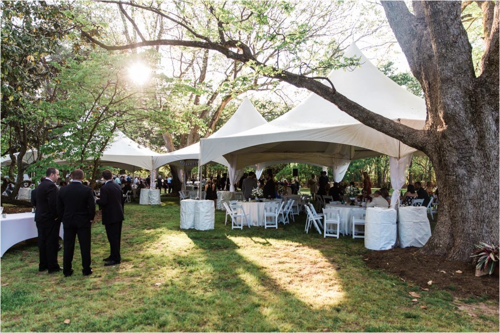 An outdoor wedding reception setup with white tent, tables and chairs under large trees at sunset.