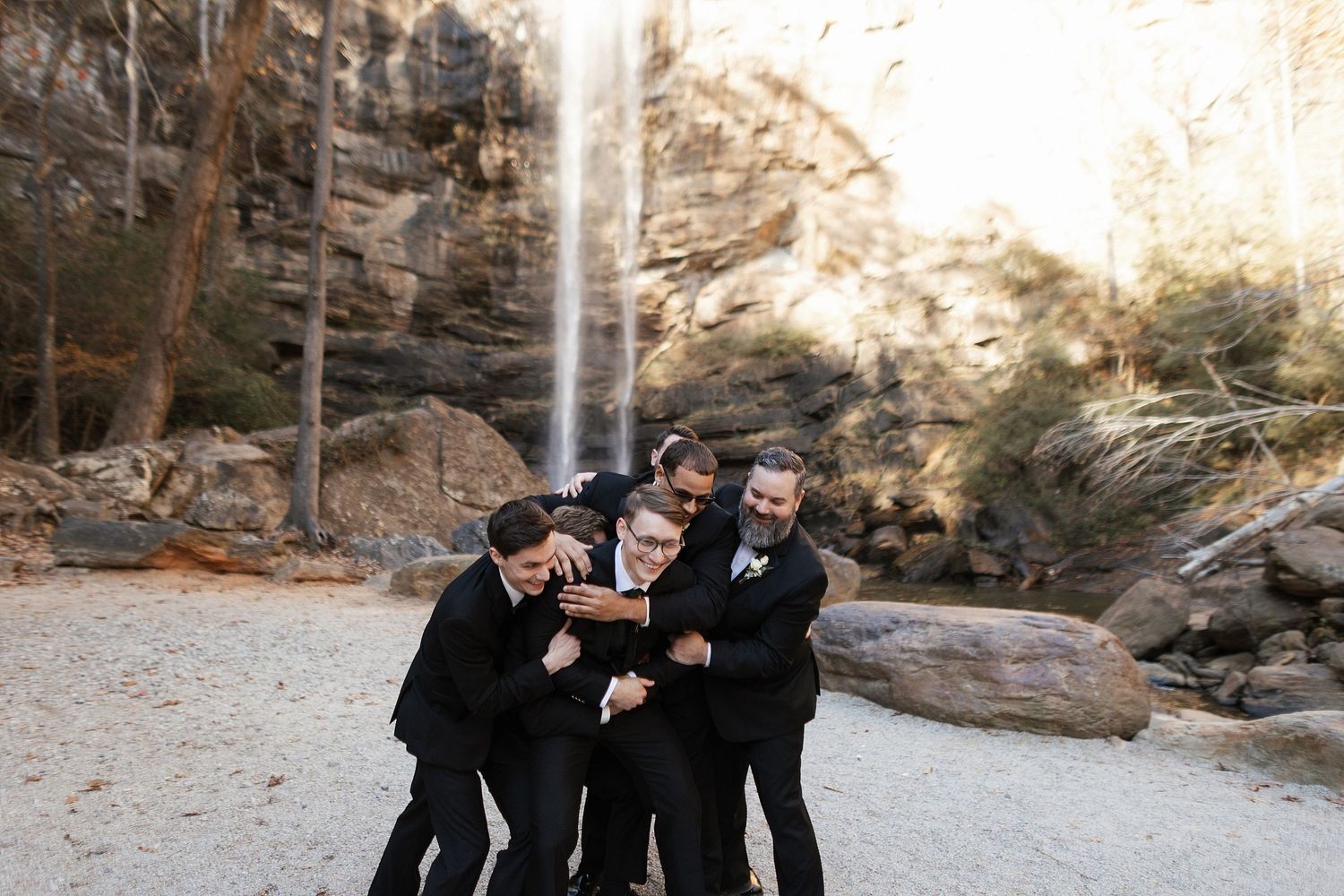 A group in black formal attire laughing and having fun together in front of a scenic waterfall.
