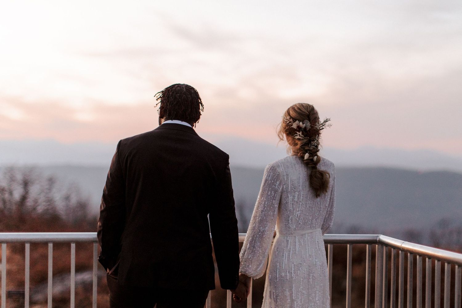 A couple in wedding attire stands at a scenic overlook against mountain silhouettes during sunset.