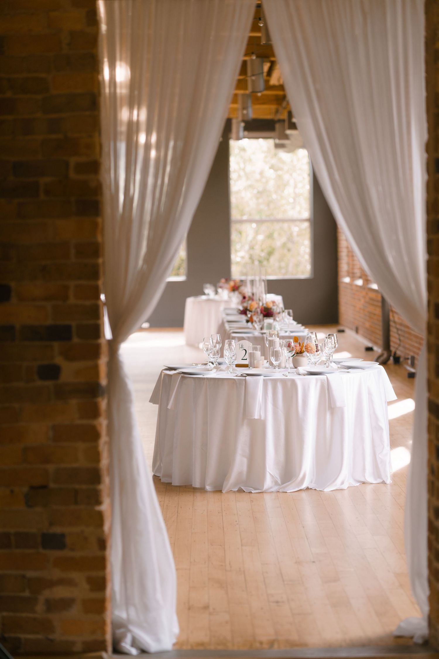 Elegant wedding reception table with white linen, floral centerpieces, and sheer draping in a rustic brick venue.