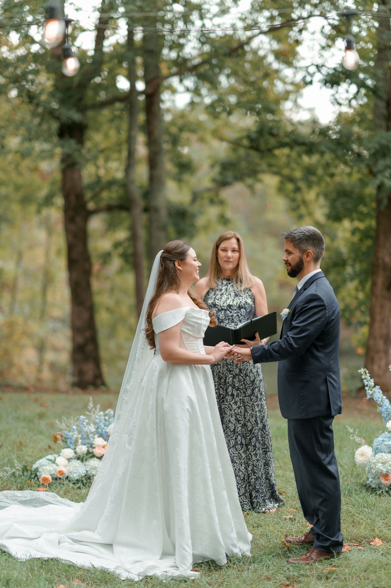 Bride and groom hold hands during outdoor woodland wedding ceremony with officiant and blue floral arrangements.