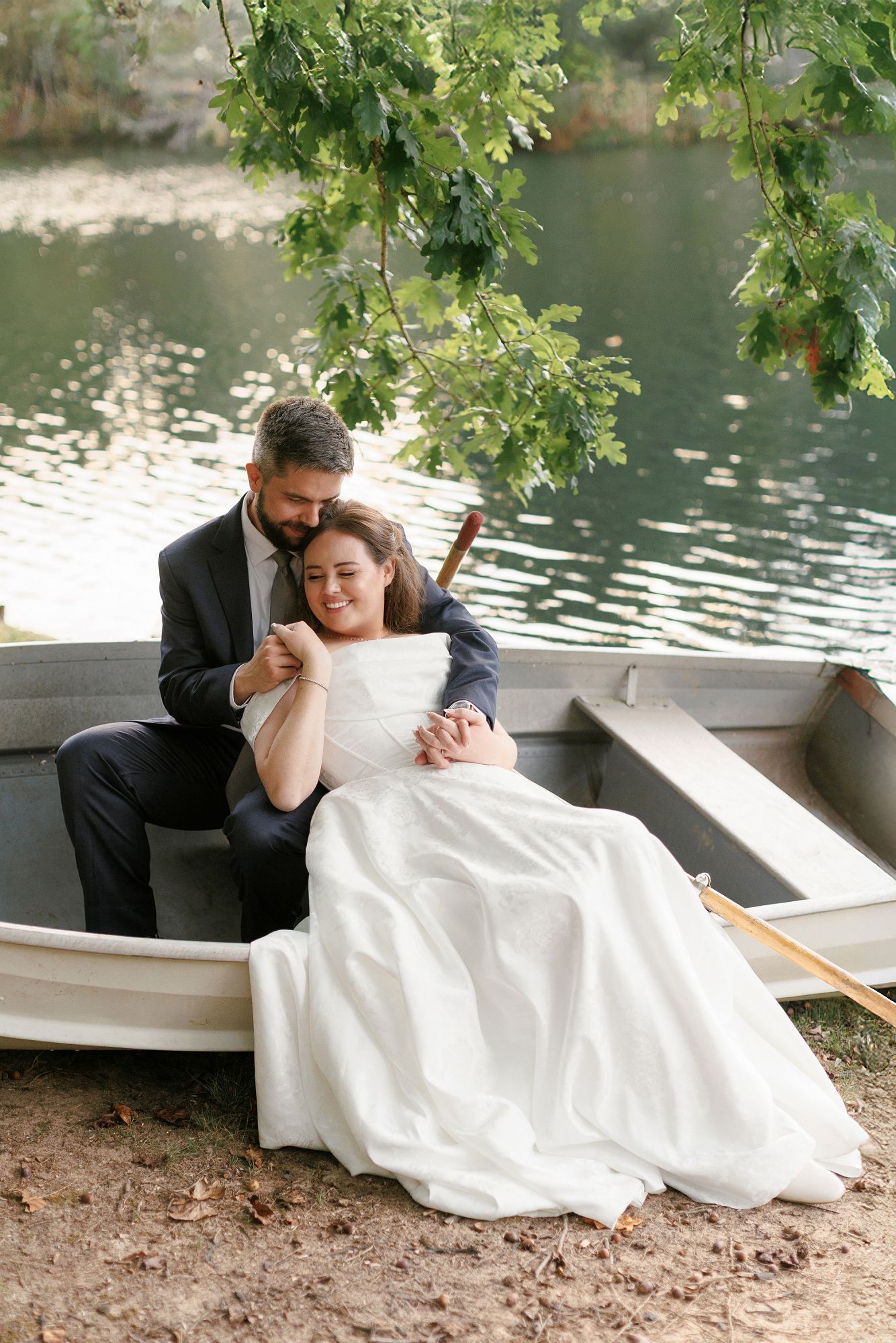 Bride and groom sitting in a rowboat by a lake, sharing a tender embrace on their wedding day.