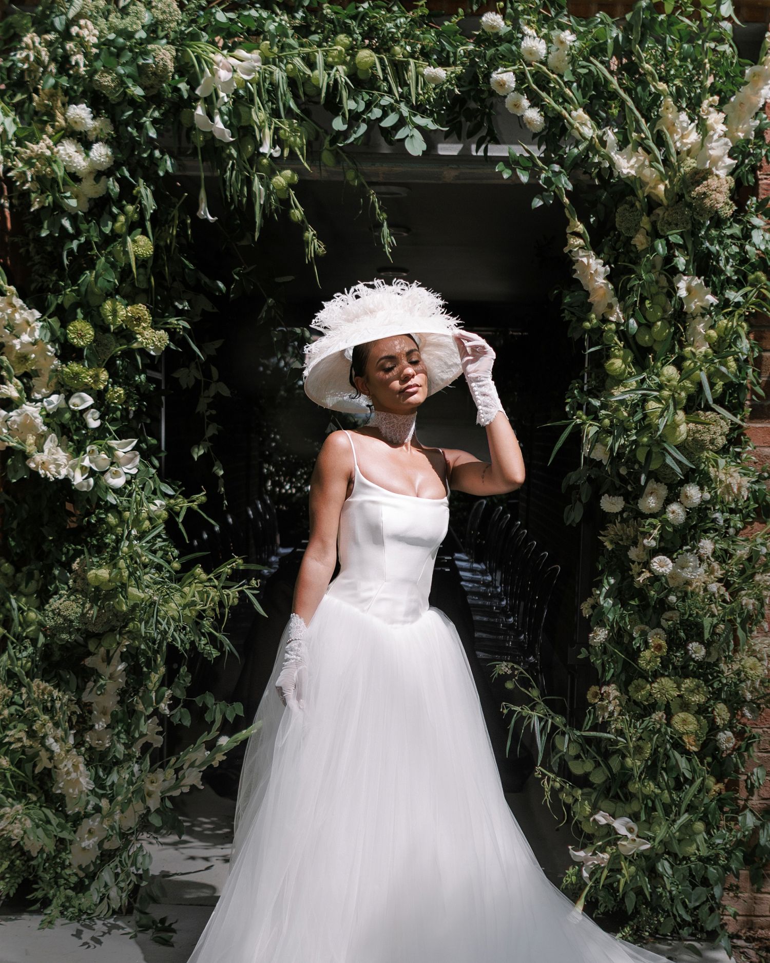 Bride in white tulle gown and wide-brim hat poses under lush floral arch with greenery and white blooms.