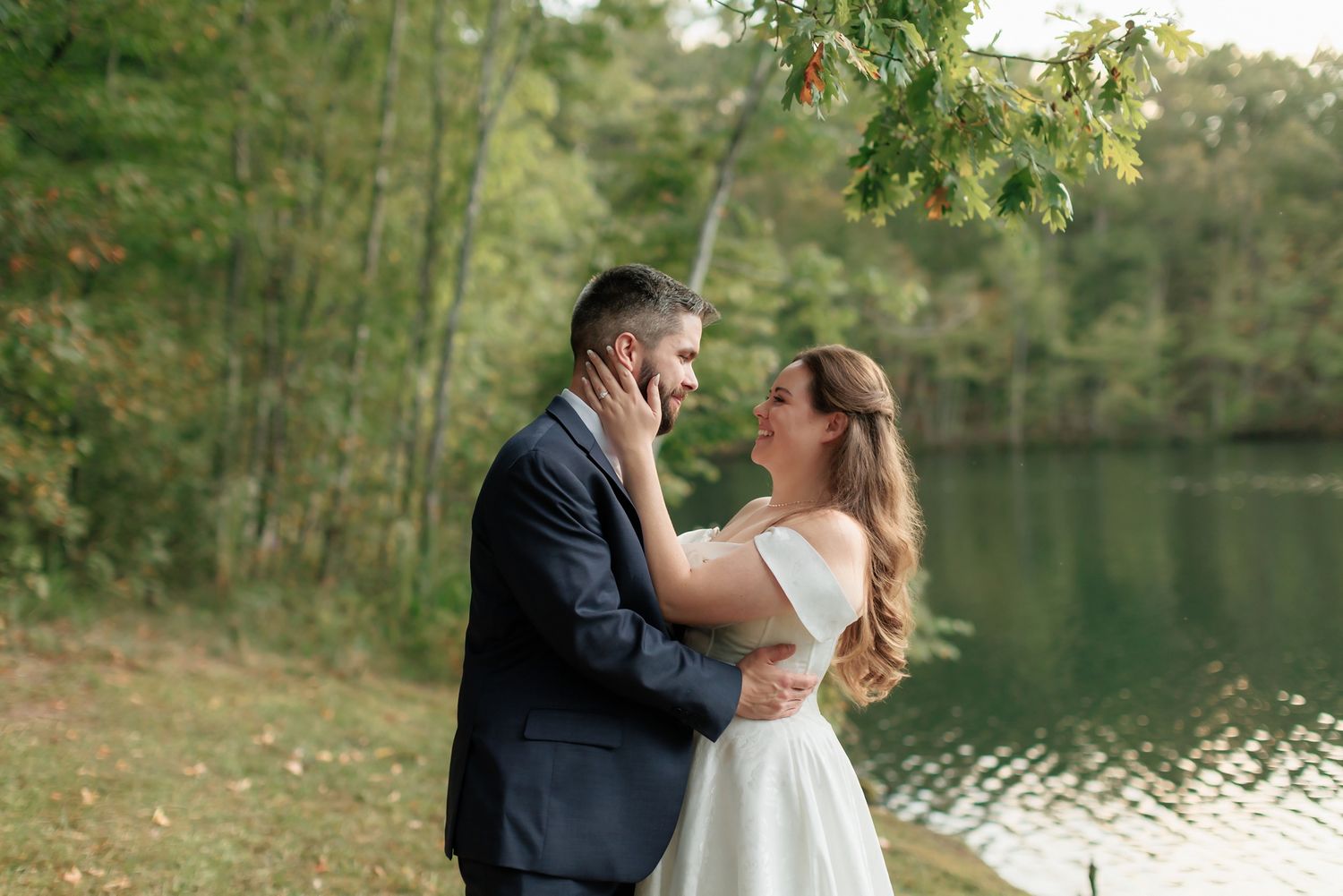 Bride and groom share a romantic embrace near a serene lake surrounded by lush green trees on their wedding day.