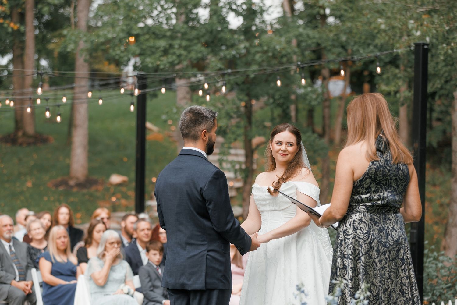 Bride smiling at groom during outdoor wedding ceremony while officiant holds book under string lights in forest.