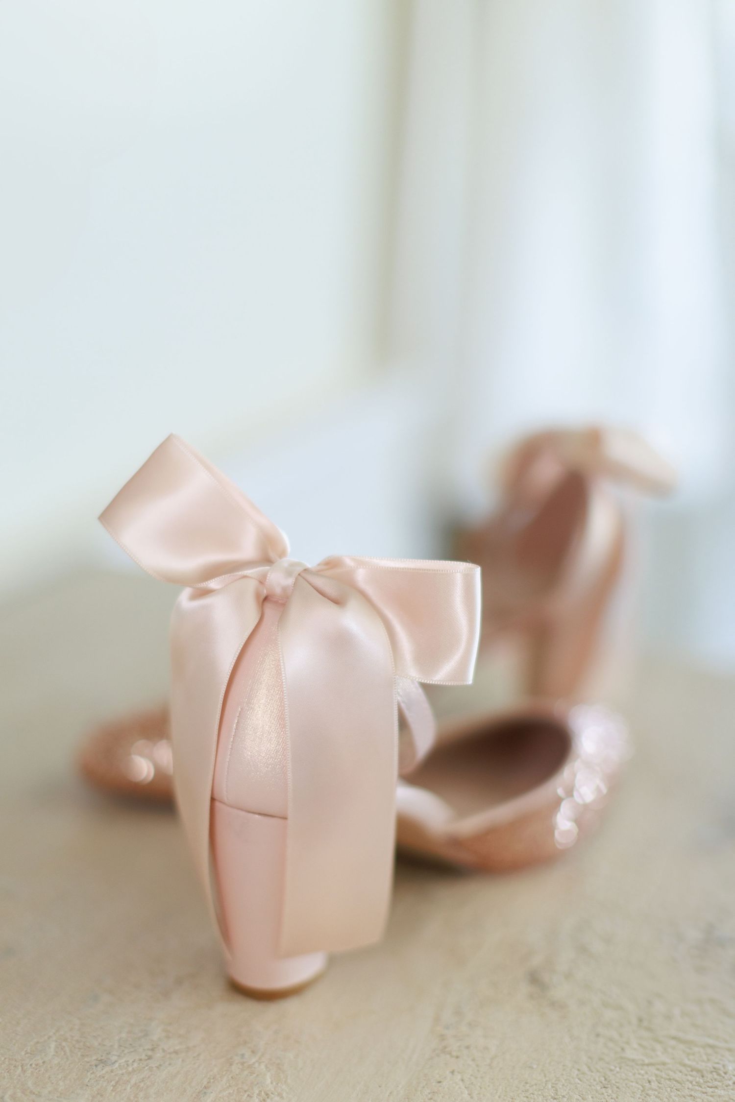 Elegant blush pink satin wedding heels with satin bow detail displayed on a soft neutral surface.
