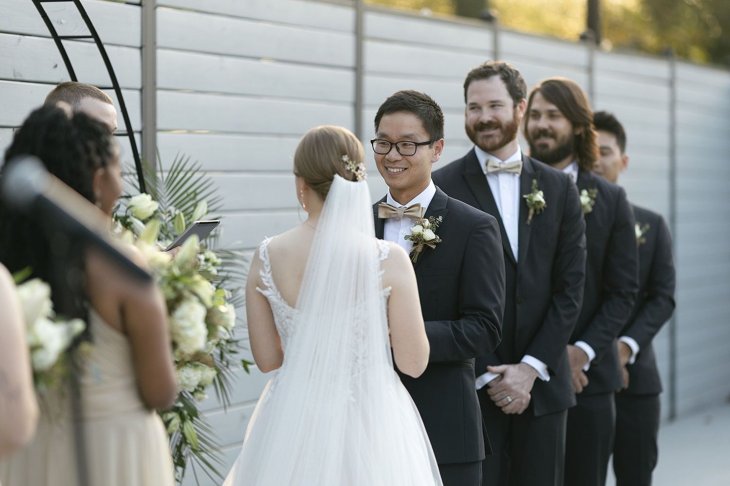 Wedding ceremony with a bride in white gown and groomsmen in black suits standing at a modern outdoor venue.