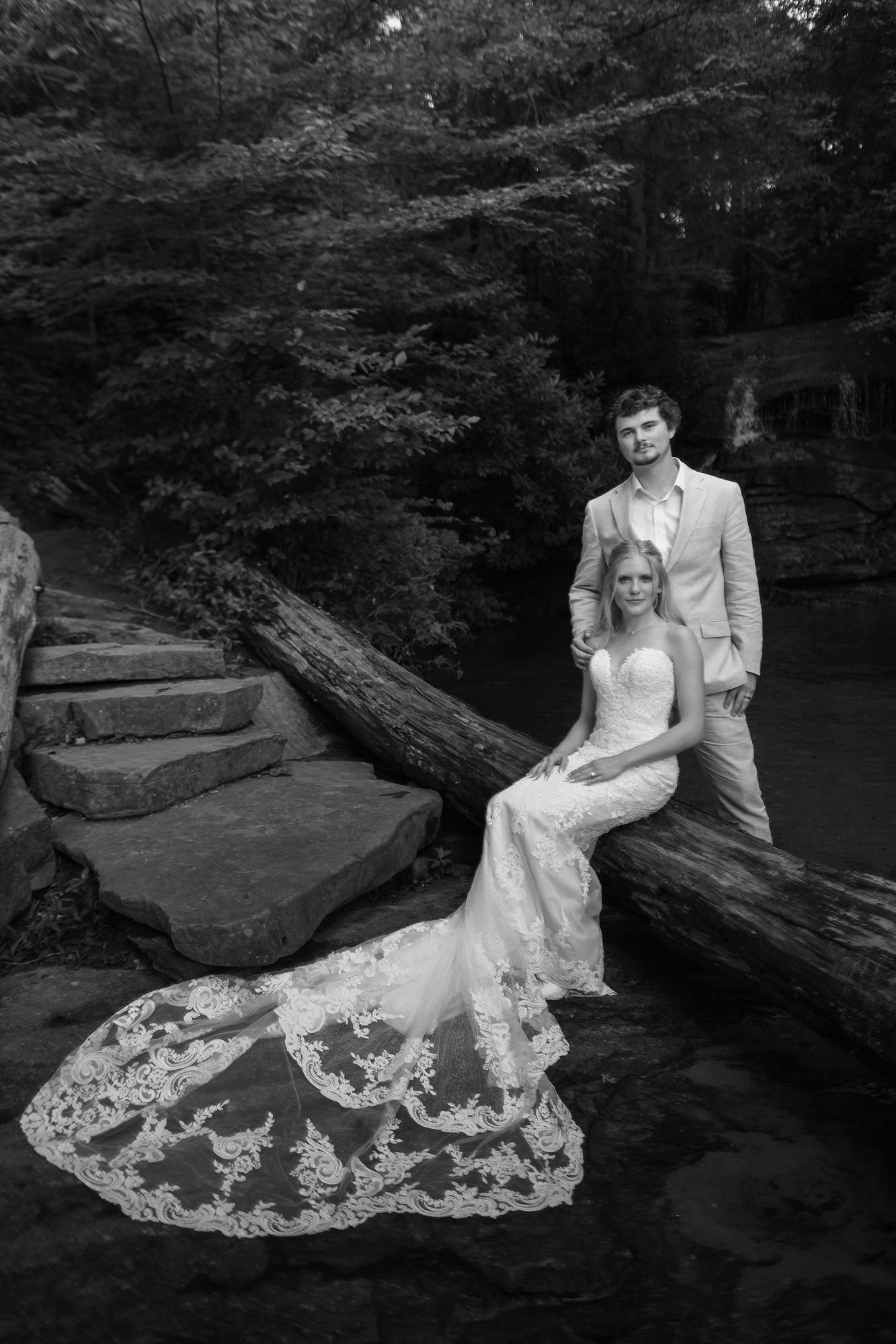 A wedding couple poses together on rustic wooden logs in a wooded setting, with the bride's lace dress beautifully displayed.