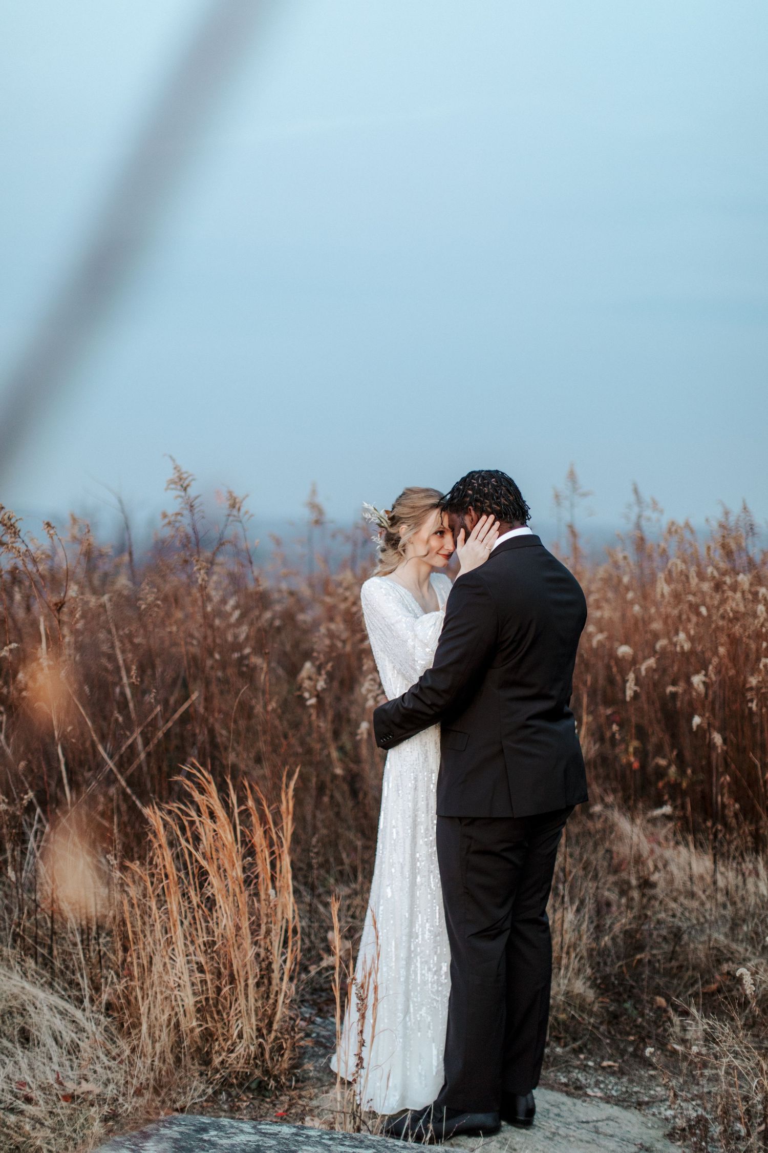 A couple in wedding attire share an intimate moment among tall dried grasses against a blue sky backdrop.