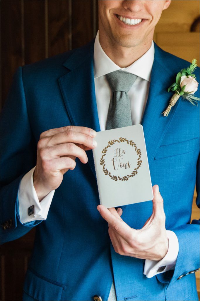 Groom in blue suit holds elegant wedding vow book with floral wreath design.