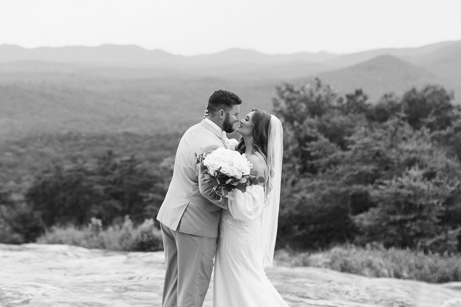 Black and white wedding portrait of couple with white bouquet against mountain landscape.