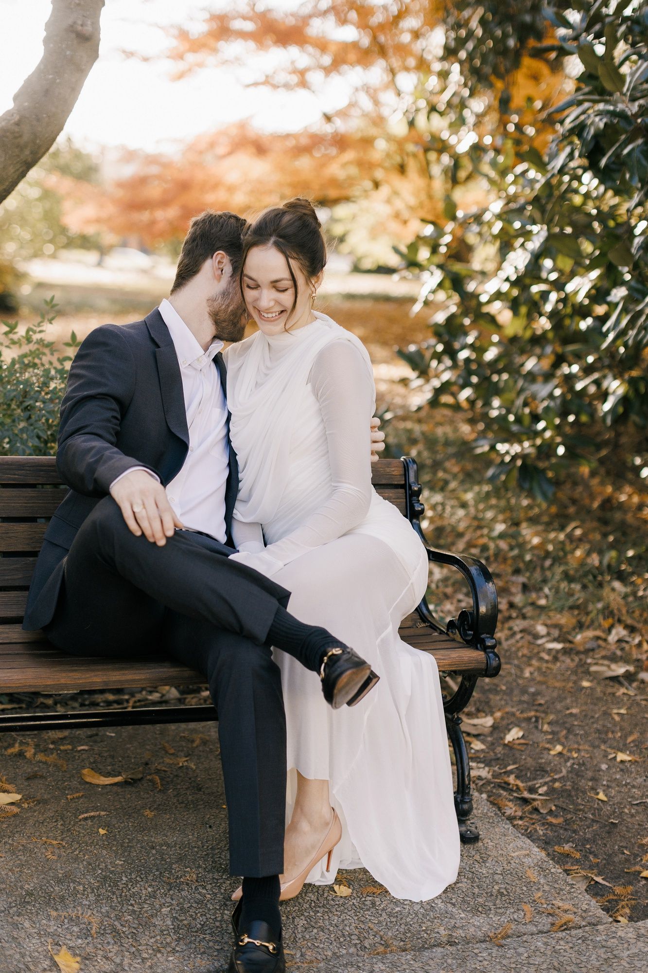 A romantic couple sits together on a park bench surrounded by autumn foliage in a natural outdoor setting.