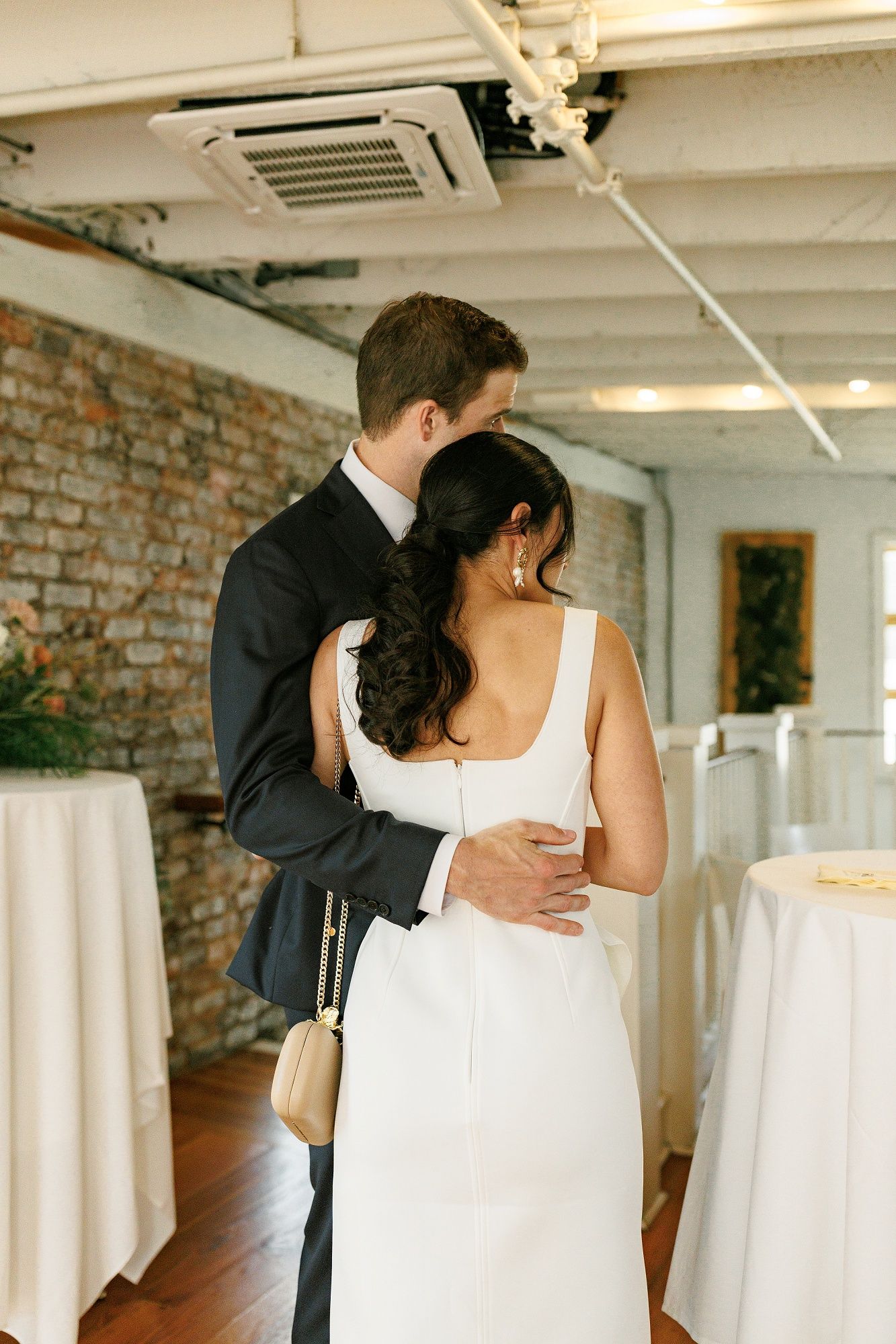 A couple embraces during their wedding reception in a rustic brick-walled venue with white linens and industrial ceiling.