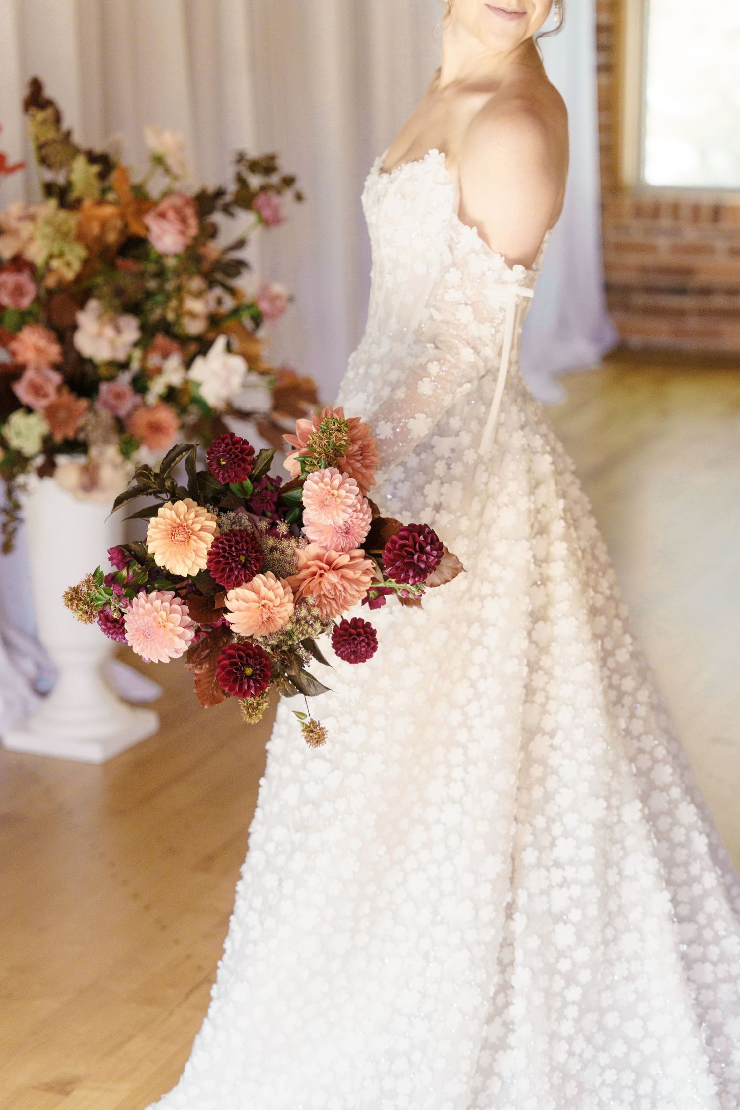 Bride in white textured ball gown holding lush pink and burgundy dahlia bouquet in elegant indoor wedding venue.