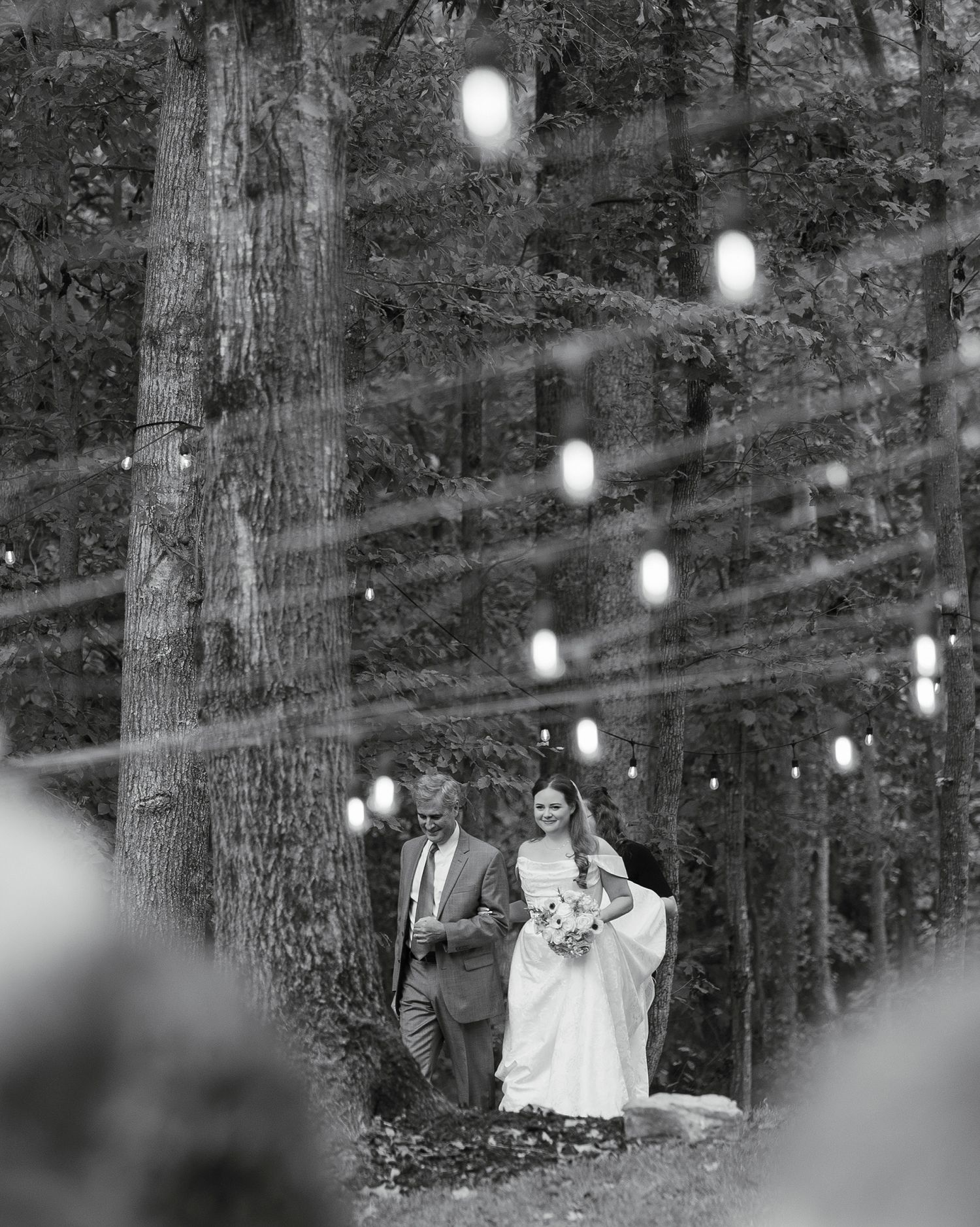 Black and white photo of bride walking down aisle with escort through tree-lined outdoor wedding venue.