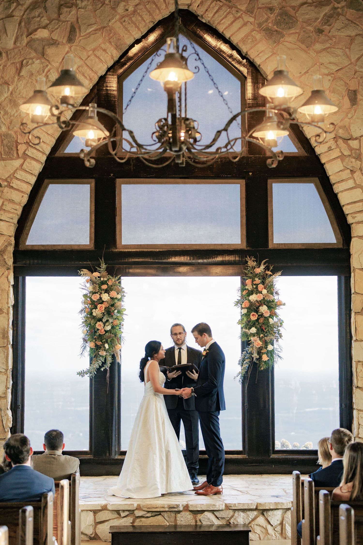Bride and groom exchange vows at stone chapel altar with floral arrangements and panoramic windows at elegant wedding ceremony.