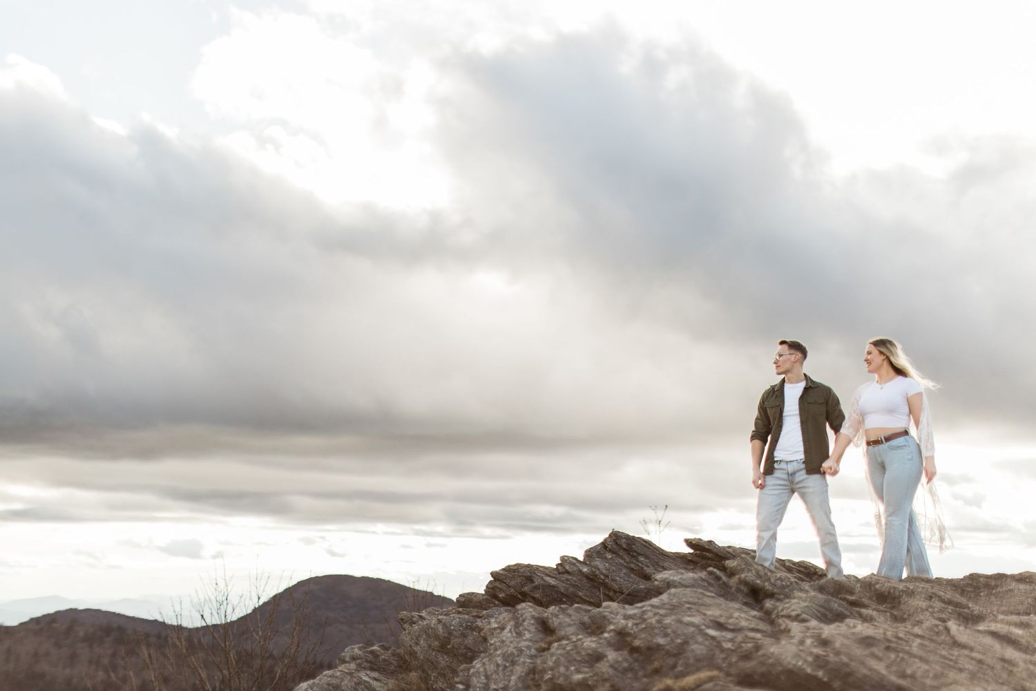 A couple stands together on a rocky mountain peak against dramatic cloudy skies.