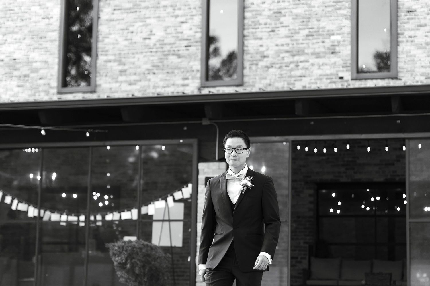 A person in a sharp black suit stands confidently in front of a brick building with string lights in the background.