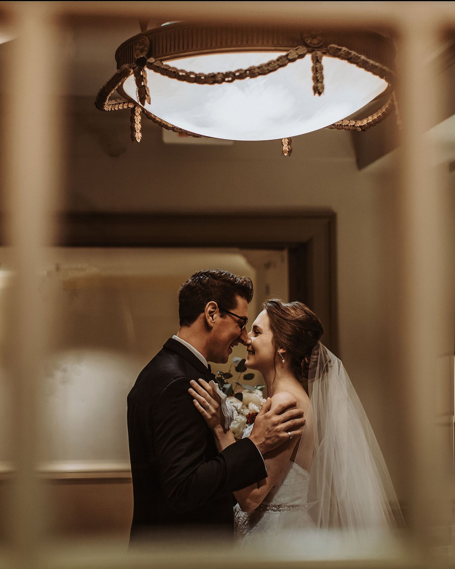 An intimate wedding moment captured through a doorframe under an ornate ceiling light fixture.
