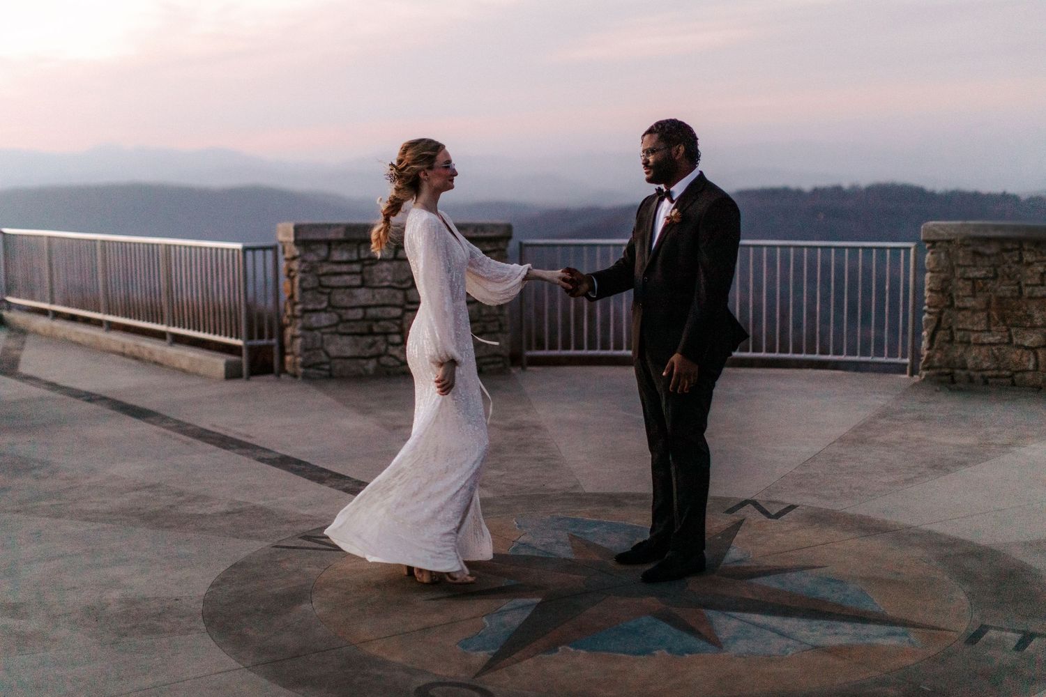 A couple shares a romantic moment at sunset on a mountain overlook with stone railings.