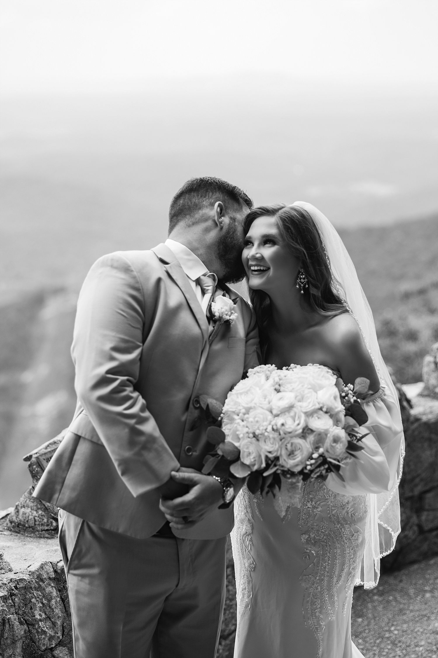 A romantic wedding couple shares a joyful moment on a mountain overlook with a white bouquet and grey suit.
