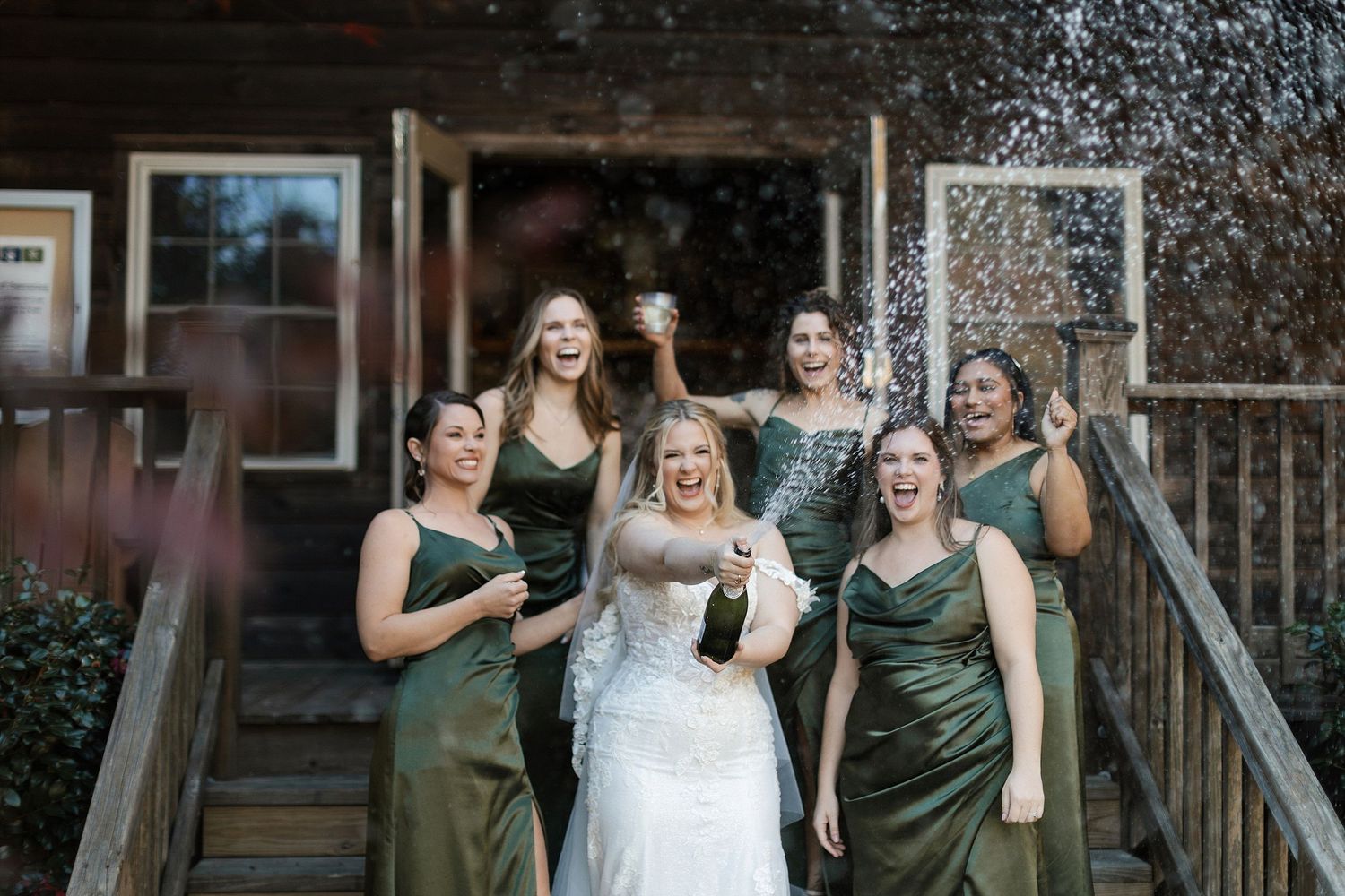Beautiful wedding party in emerald green bridesmaid dresses pose and laugh together on rustic wooden steps.