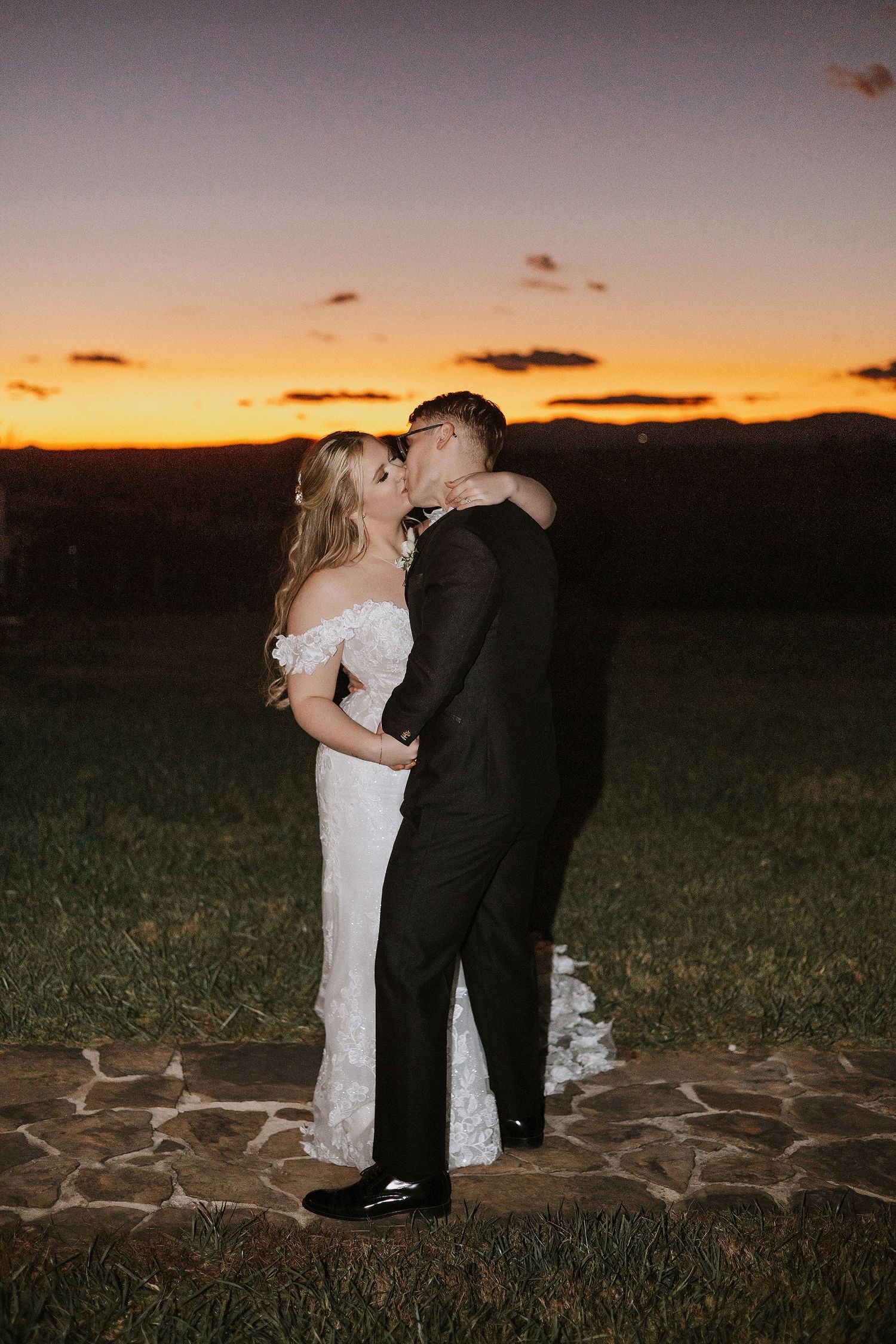 A couple in wedding attire share a romantic kiss at sunset with mountains in the background.