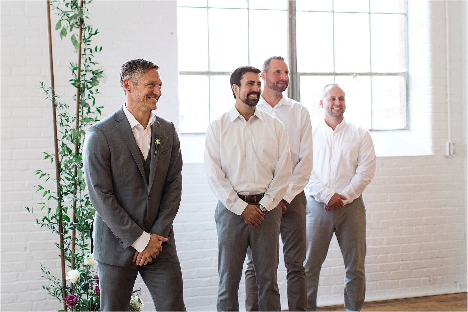 Groomsmen in light colored suits share a candid moment in a bright white venue with greenery.