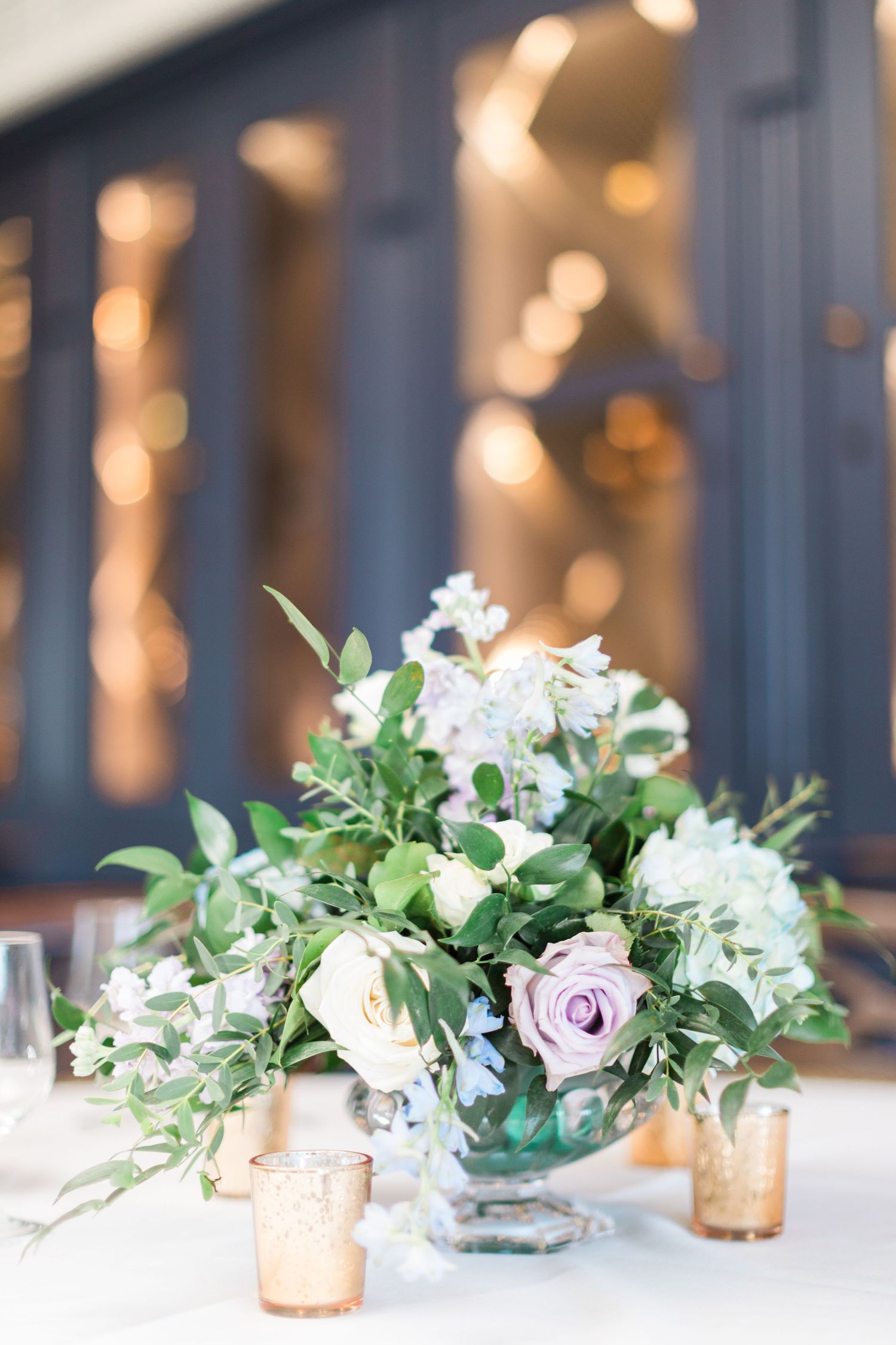 Elegant wedding centerpiece with white and lavender roses, greenery in glass vase surrounded by gold votive candles.