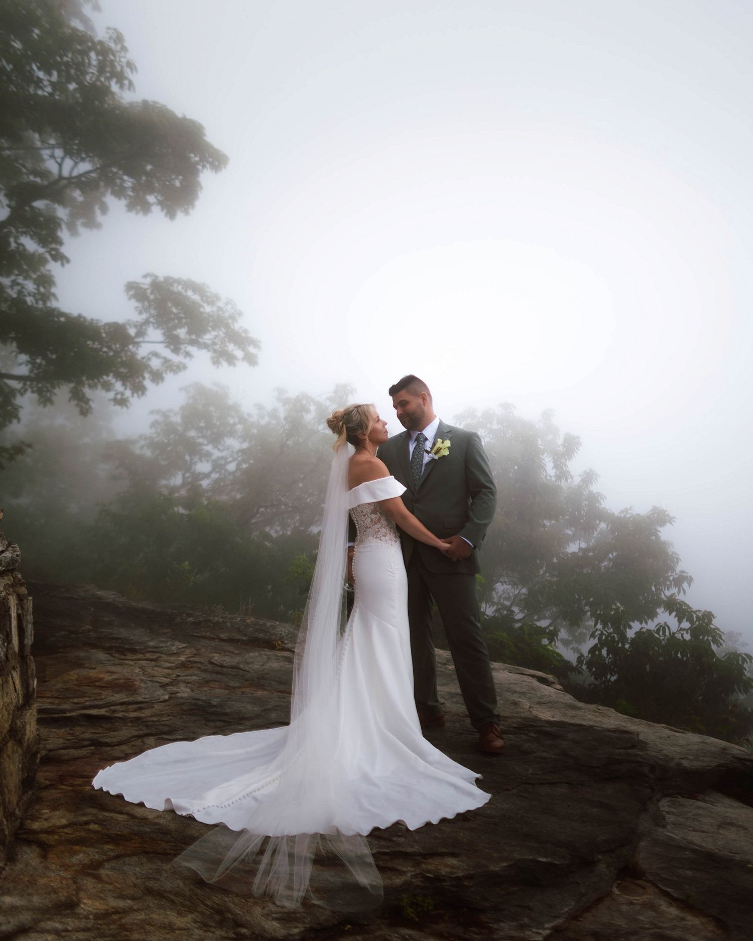 Bride and groom standing on rocky misty mountaintop, facing each other holding hands in wedding attire.