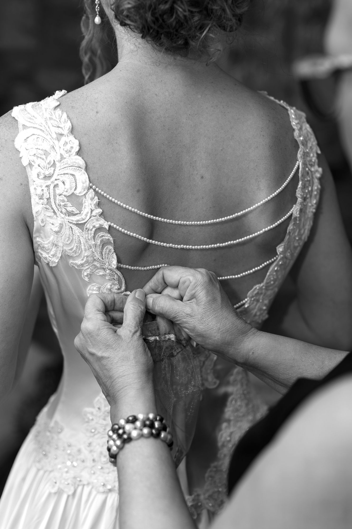 Black and white photo of a bride having her lace wedding dress fastened, featuring elegant chain back detail.