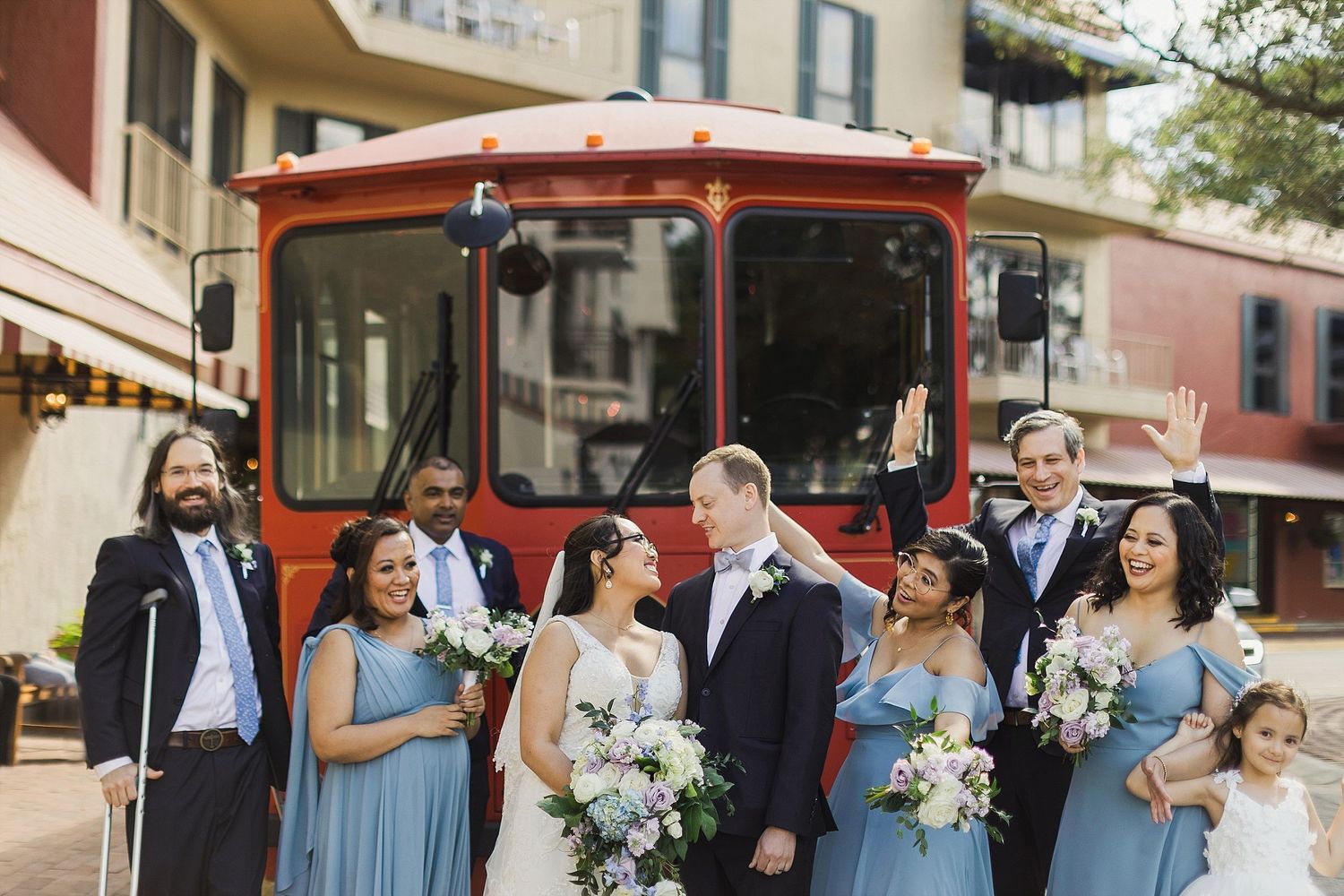 Wedding party in blue dresses and suits posing in front of a red trolley on a city street.