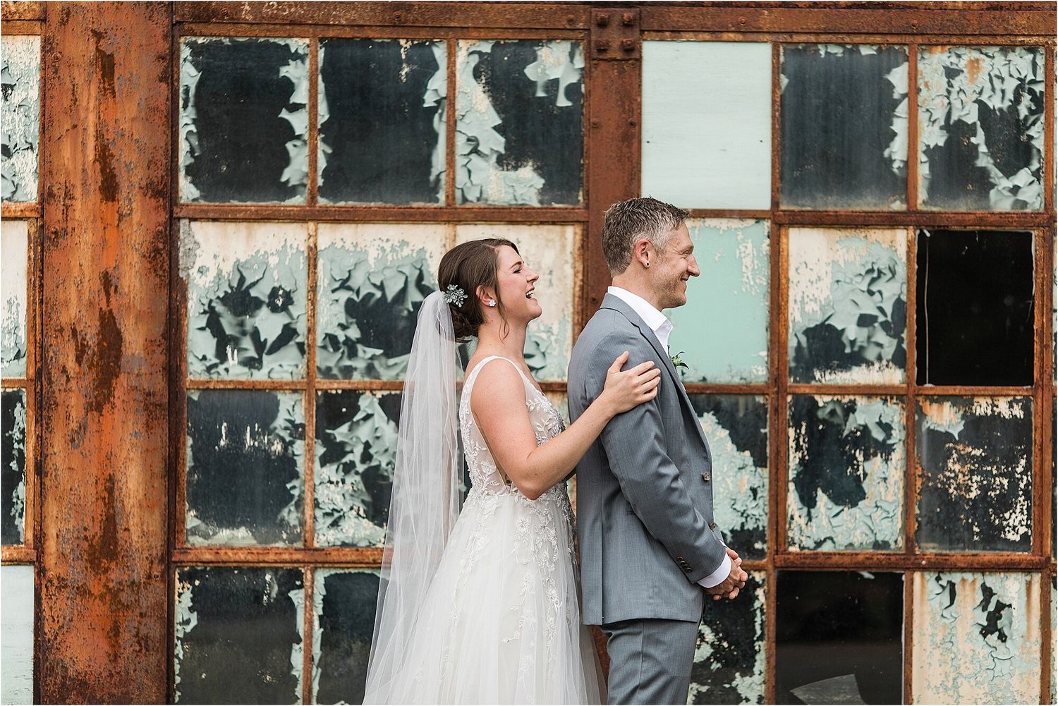 Couple in wedding attire standing against weathered industrial window backdrop.