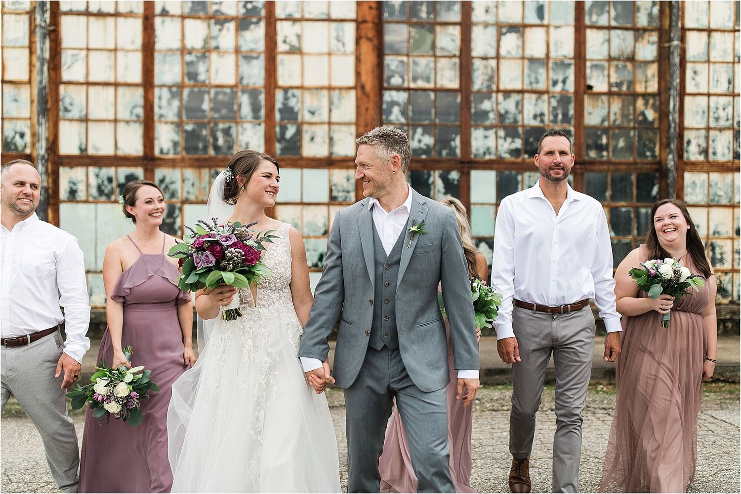 Wedding party laughs together in front of industrial backdrop with vintage windows.