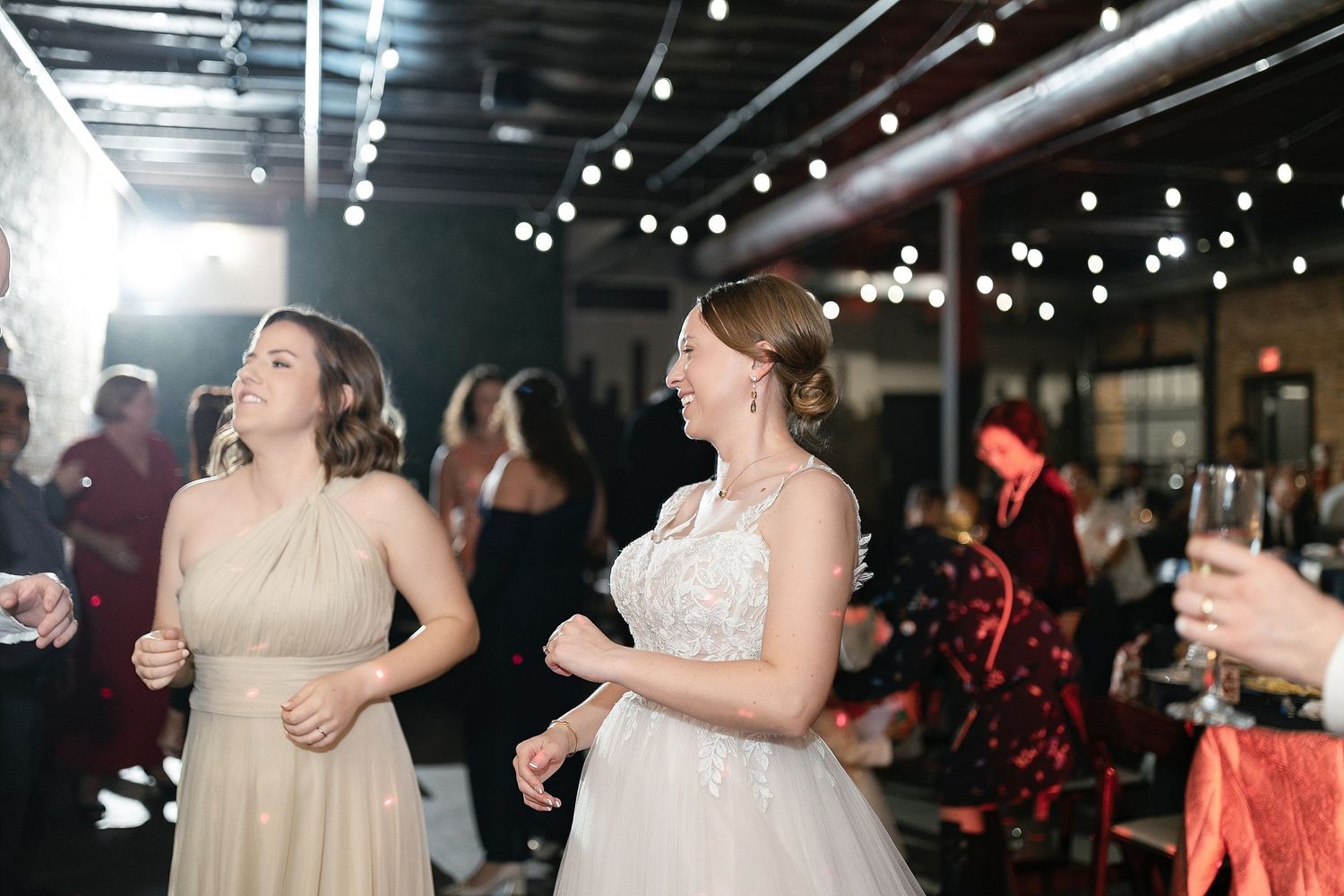 Wedding guests in formal attire dancing together on a dark dance floor with spotlights overhead at an indoor reception.
