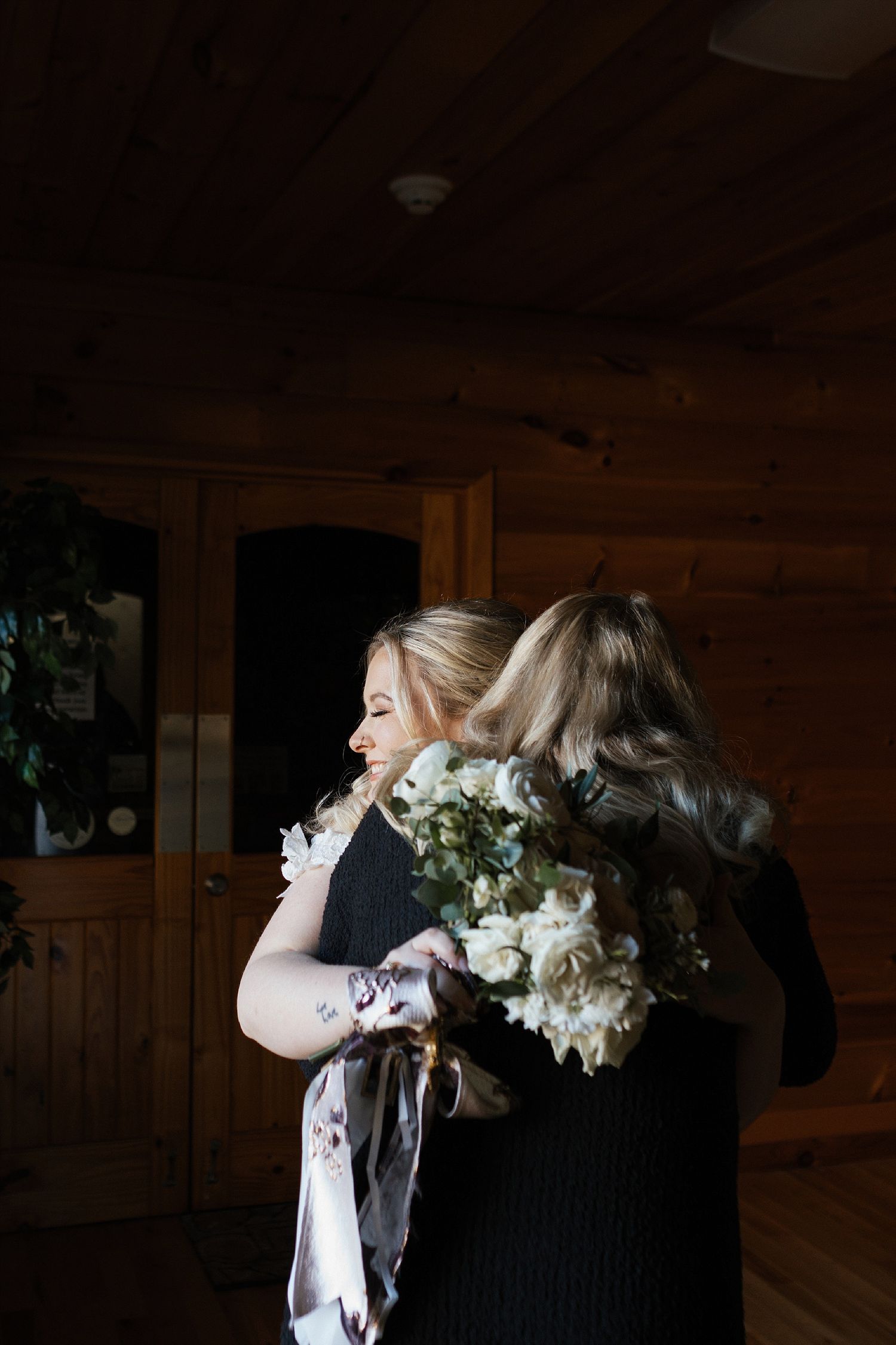 Two people share a tender embrace while holding a bouquet of white flowers in a rustic wooden room.
