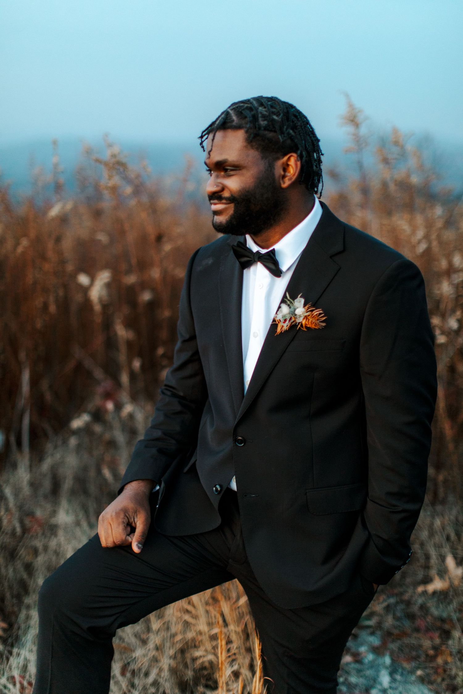 A groom in formal attire smiles while posing outdoors against natural scenery.