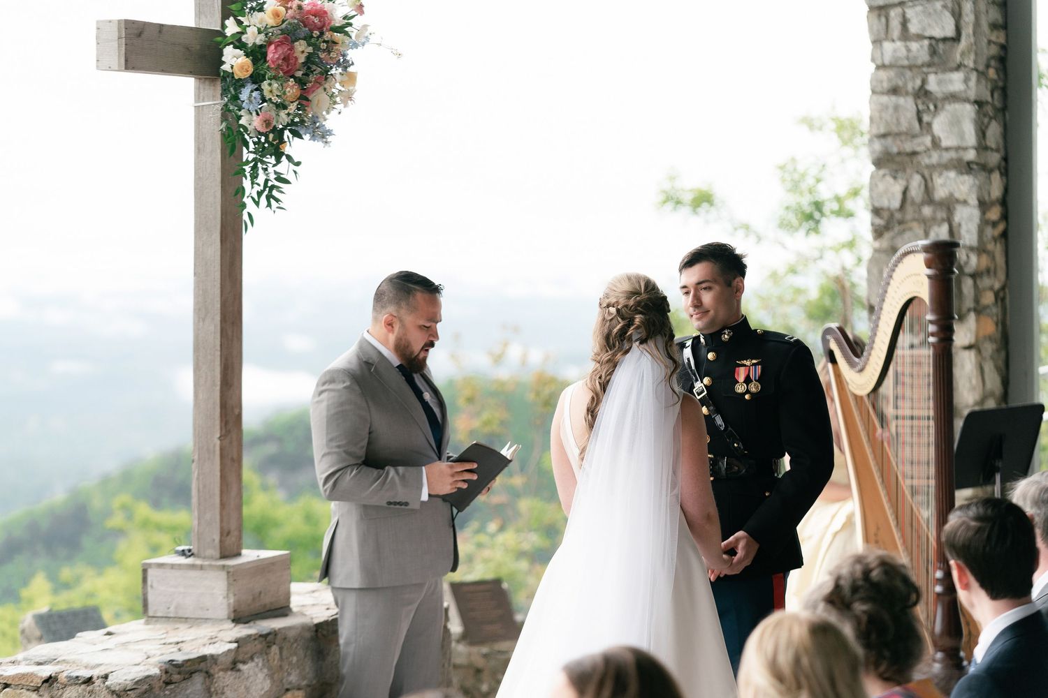 An outdoor mountain wedding ceremony takes place under a wooden pergola decorated with pink florals and a harp nearby.
