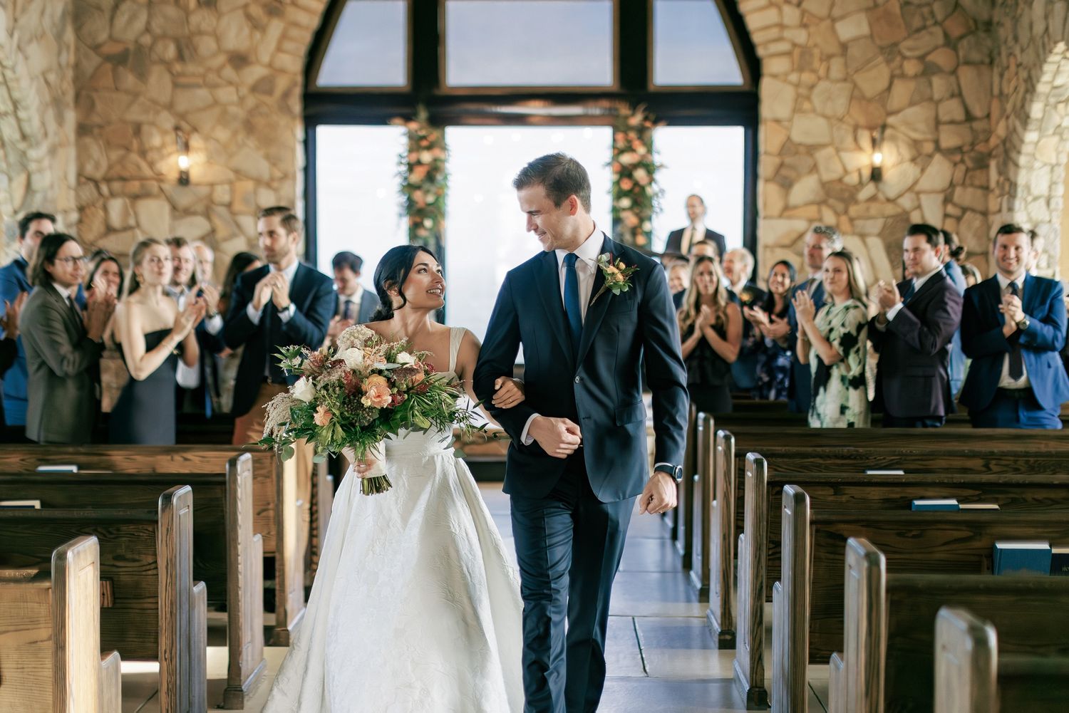 Wedding party stands in a rustic church with exposed brick walls and wooden pews during an emotional ceremony.