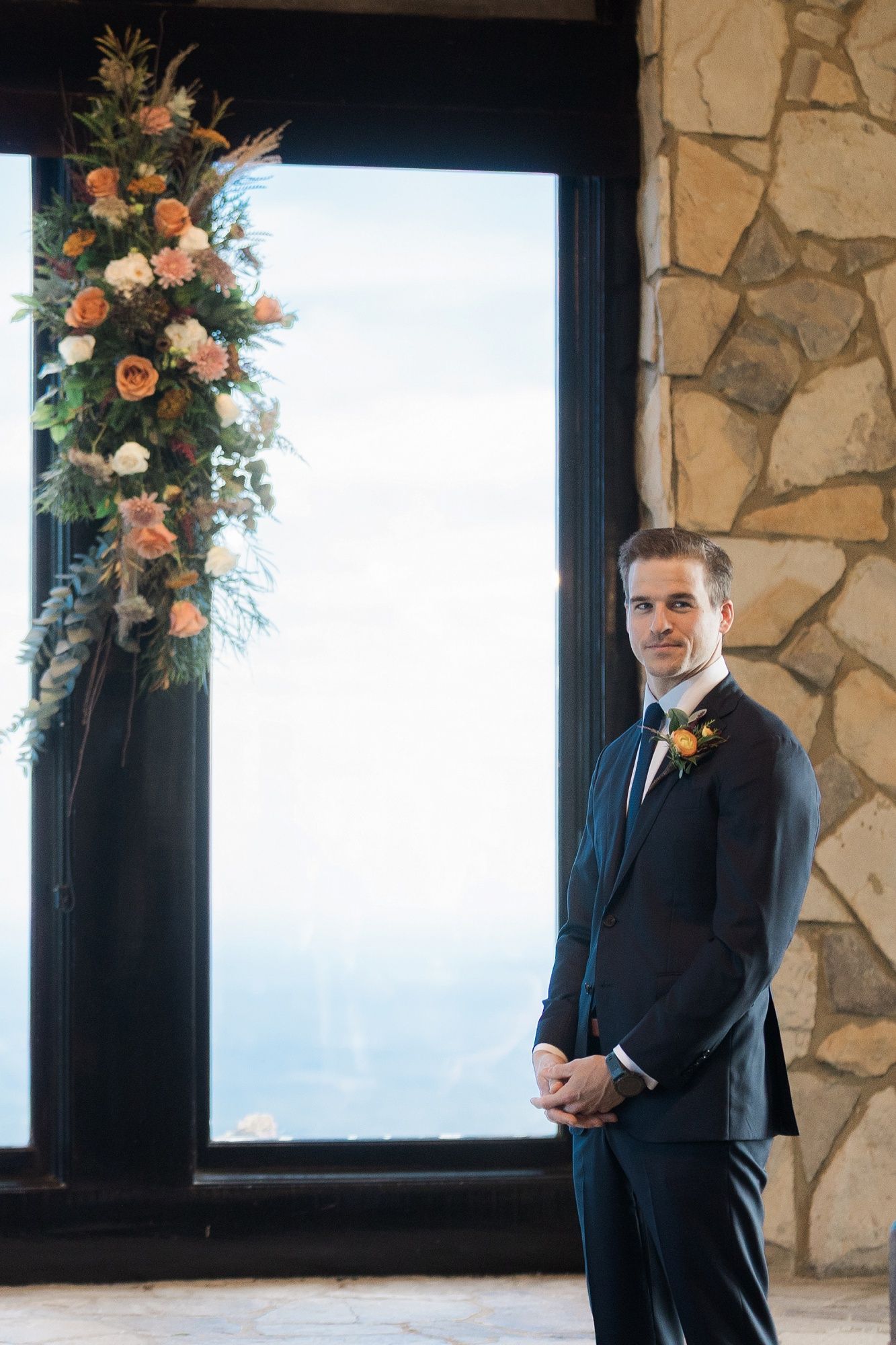 Groom in a dark suit stands near stone wall and large windows with floral arrangements at a mountain venue.
