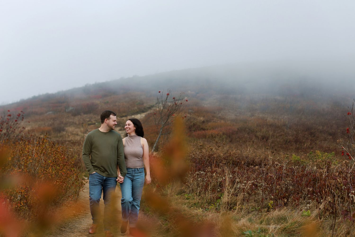 A couple walks through a misty mountain landscape with autumn foliage and fog rolling across the terrain.