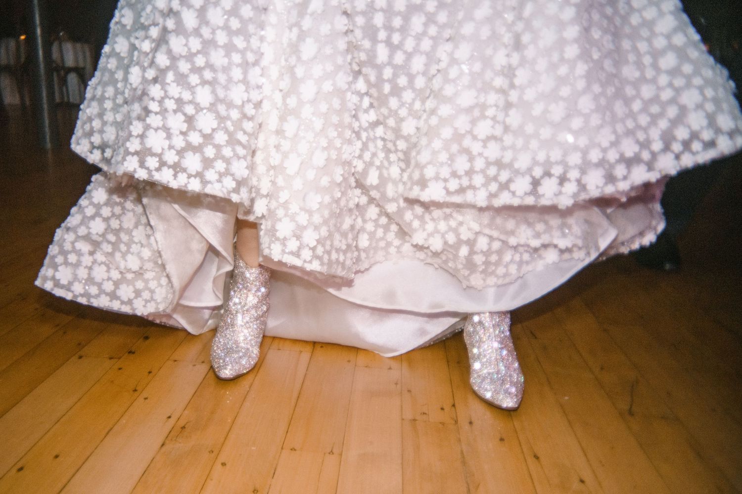 Close-up of bride's embellished heels peeking beneath a floral appliqué wedding ball gown on a wooden dance floor.
