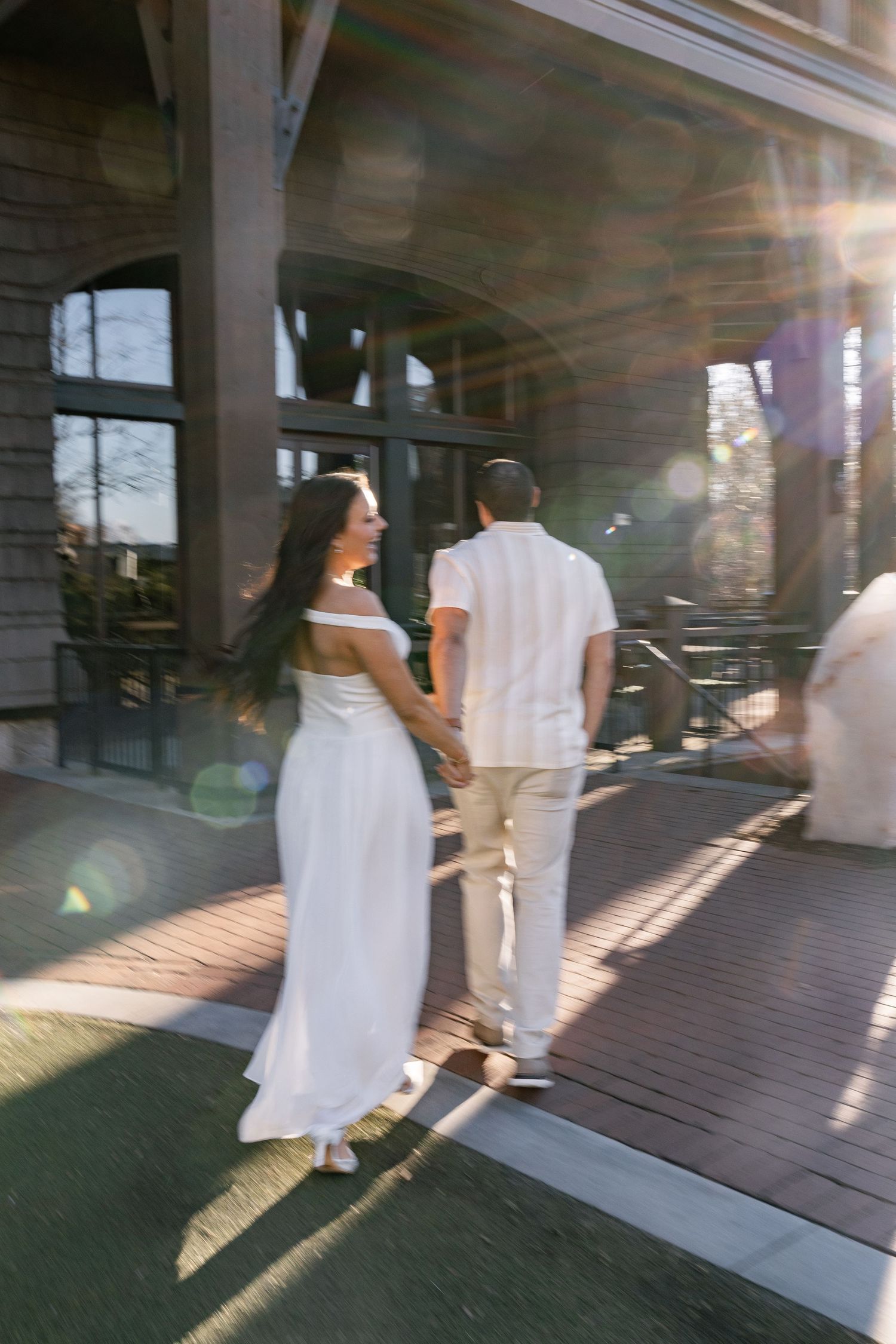A couple in white attire walks across a wooden deck as sunlight streams through creating dramatic shadows.