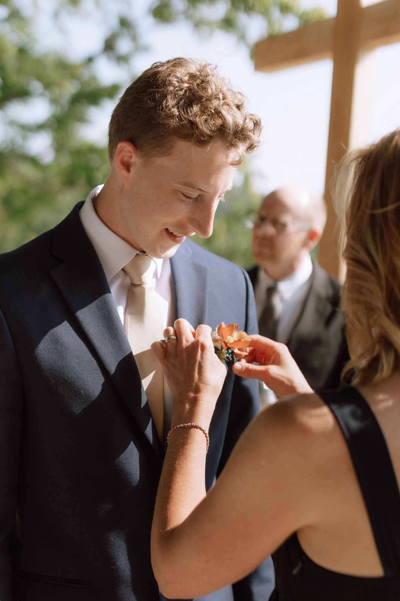 Smiling groom in navy suit has boutonniere pinned to lapel before outdoor mountain wedding ceremony.