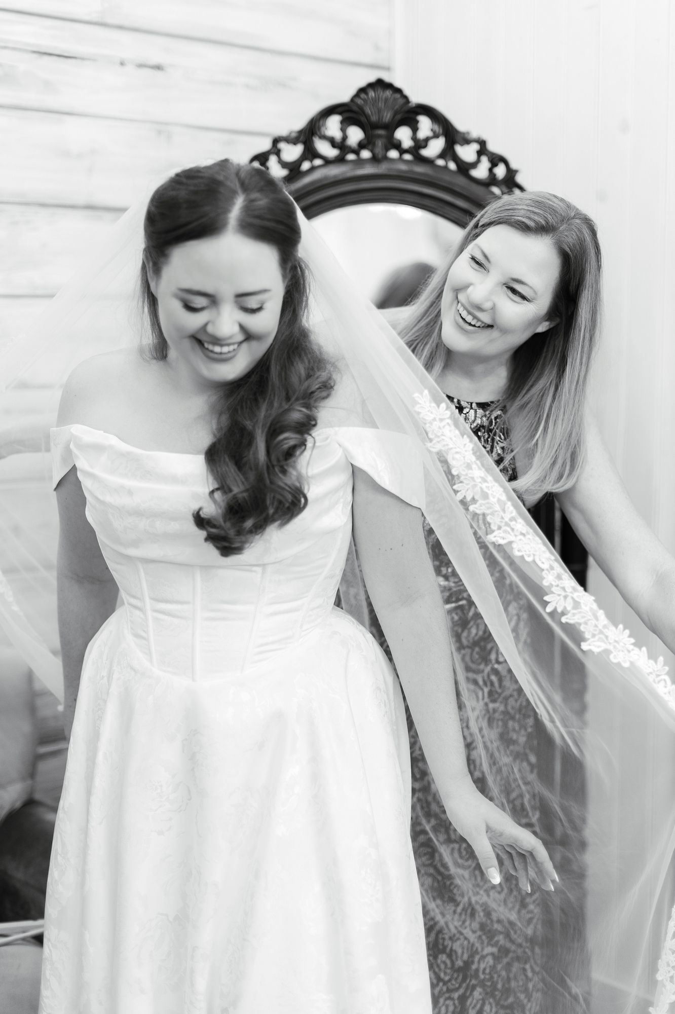Black and white photo of a smiling bride in an off-shoulder gown as her bridesmaid adjusts her lace veil.